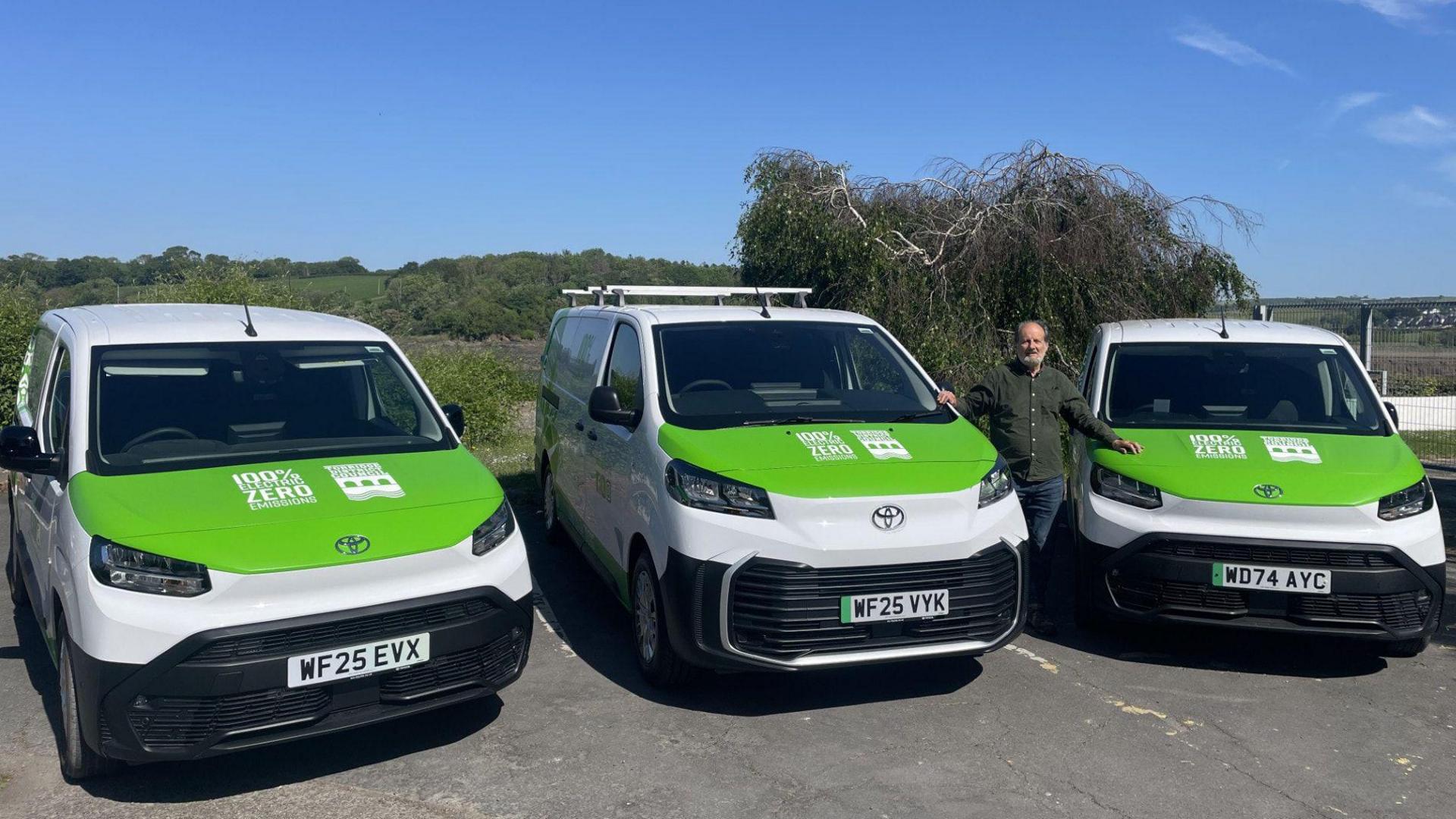 Councillor Peter Hames standing with three electric vans that are white with green bonnets.