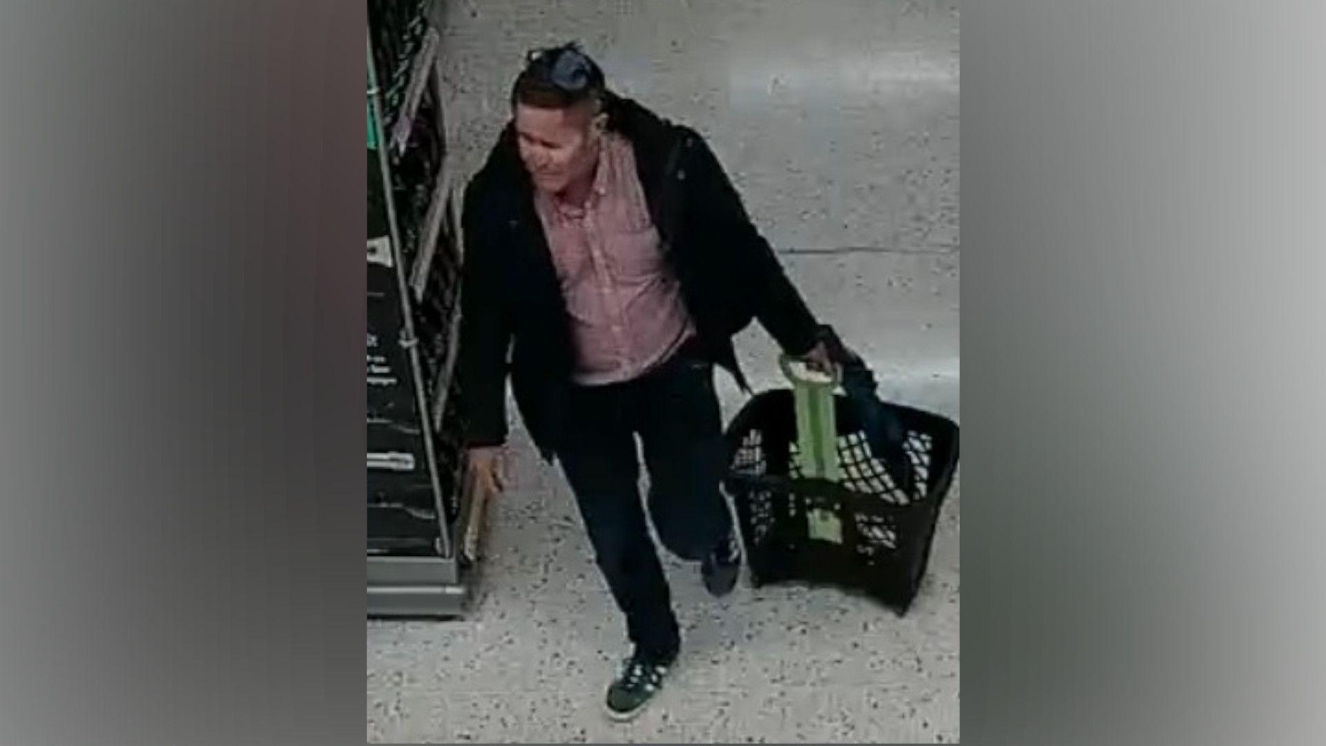 Robert Brown with dark hair wearing a dark jacket and trousers and pink shirt carrying a black plastic shopping basket and walking past some black crates in a supermarket