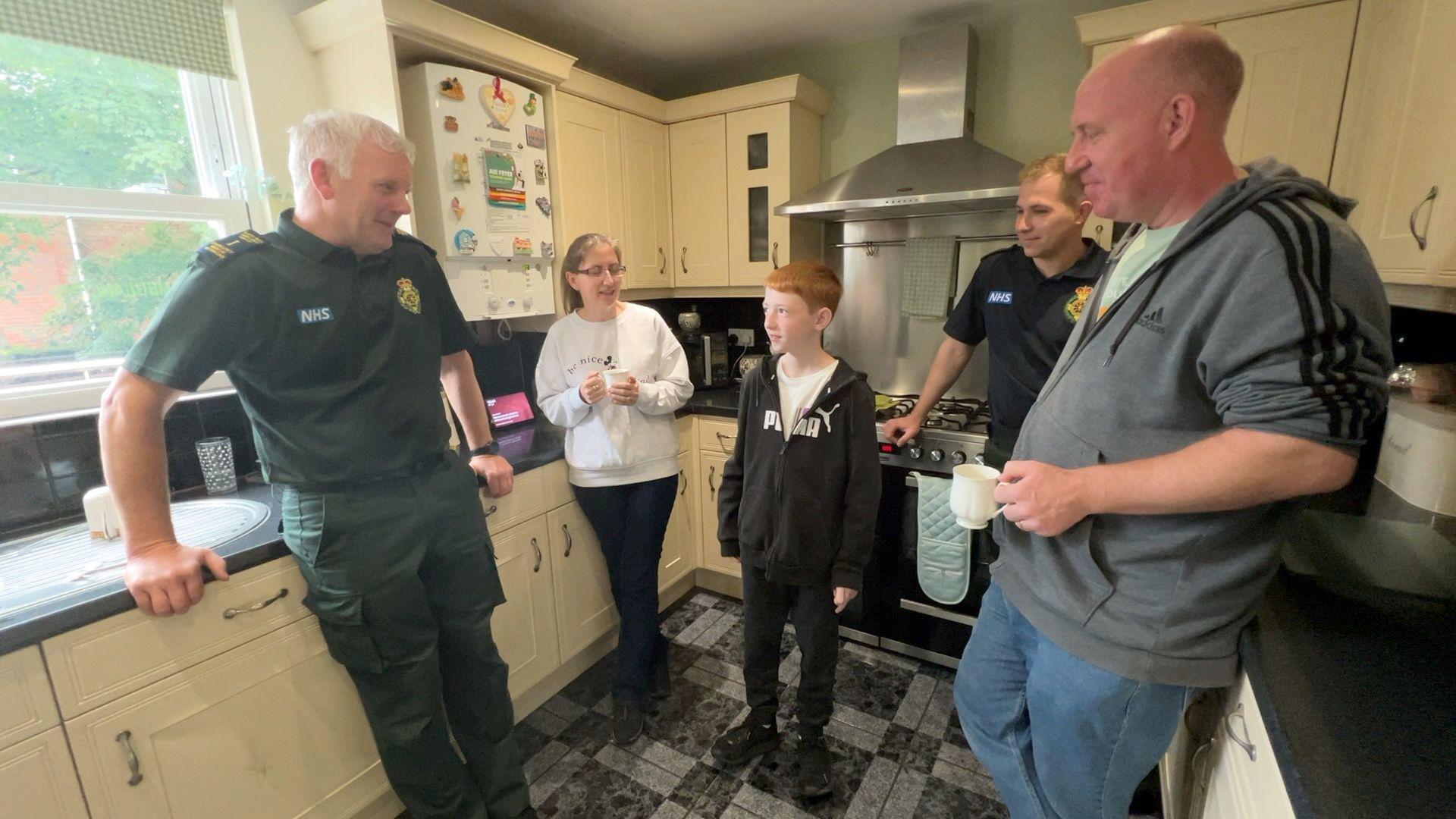 Mr Palin in a paramedic's uniform, with two men, a woman, and 11-year-old Matthew, who are all standing in a cream-coloured kitchen looking at Matthew