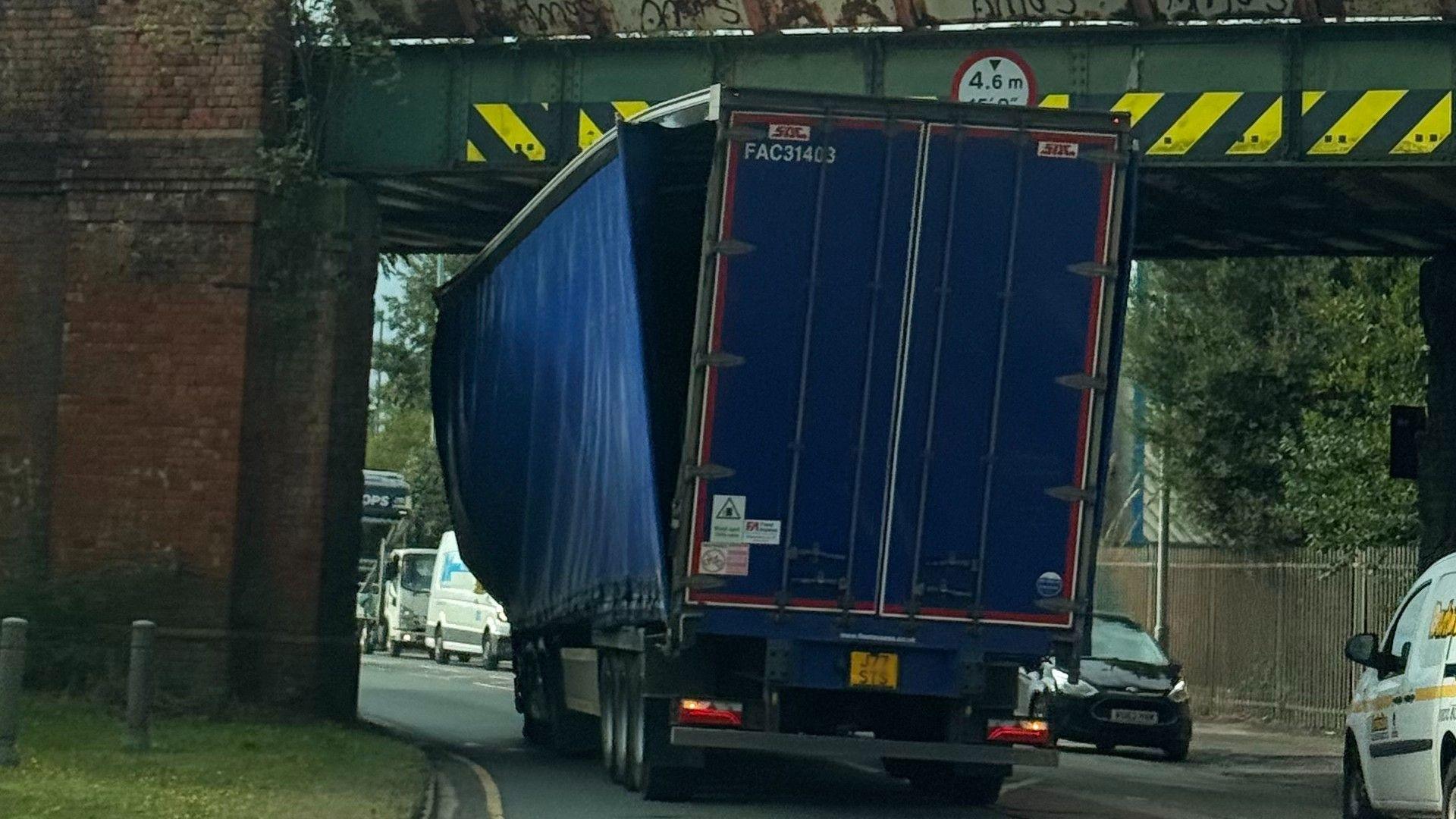 A lorry stuck under a railway bridge - there are yellow chevron markings at the top of the bridge - and the curtain on one side of the lorry is hanging off, with visible damage to the top of the vehicle.