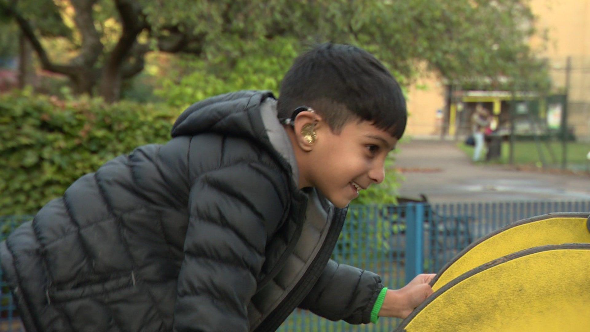 Muhaymin, a young boy wearing a black and grey coat and using a gold glittery hearing aid, climbs up a yellow slide in a children's play area