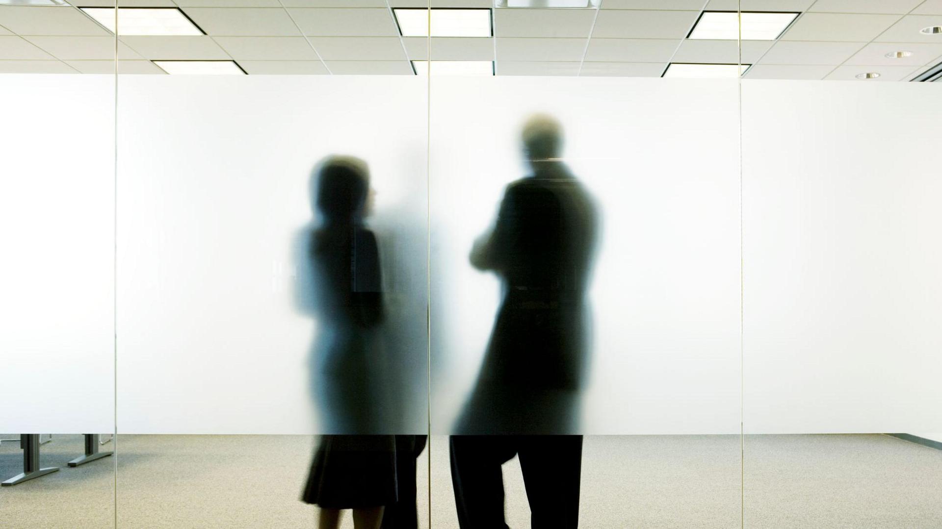 Generic image of three colleagues standing behind frosted glass in office, talking. Their faces can't be seen. The woman is wearing a black dress and the man is wearing a black suit. 