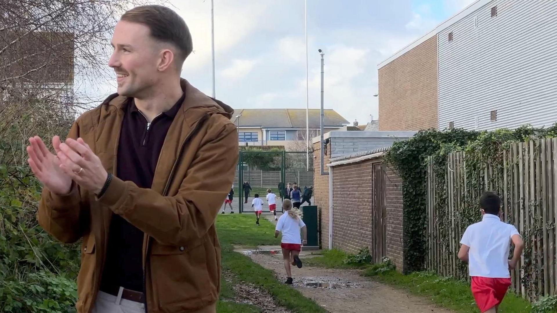 Chris is wearing a brown jacket and dark shirt is standing on a dirt path lined with greenery and wooden fencing, clapping hands. In the background, several children in white shirts and red shorts are running along the path toward a sports field. The scene appears to be part of an outdoor activity or race near school buildings under a partly cloudy sky.
