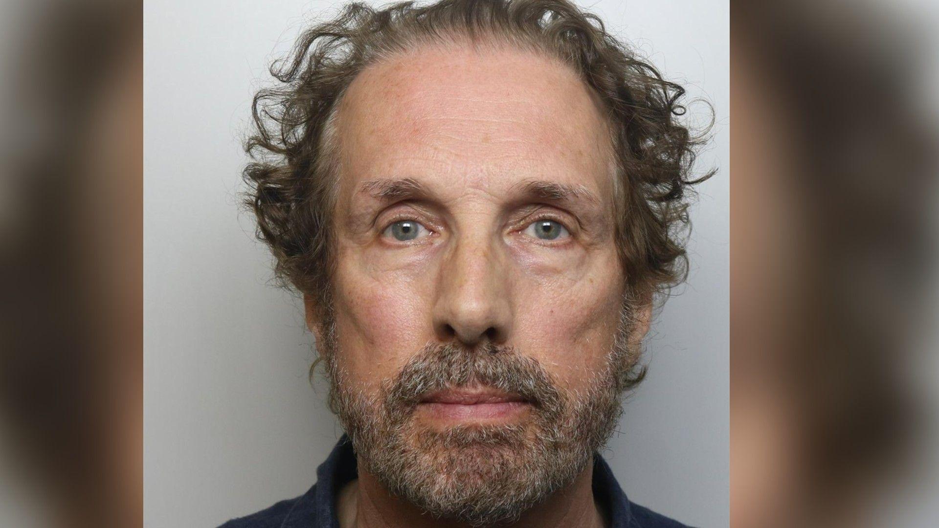 A police mugshot of Chris Brain. He has curly, swept back brown hair and a grey and brown-flecked beard.