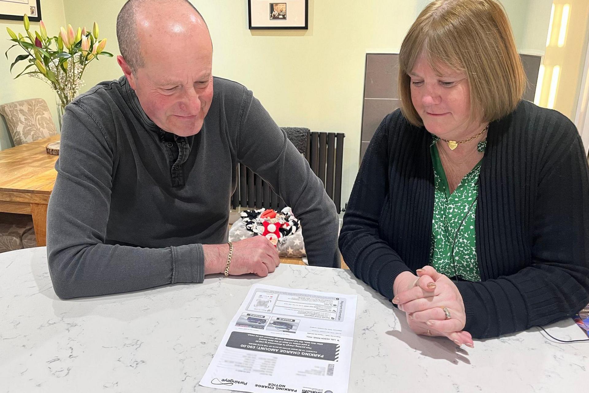 A man and a woman sit at a marble-topped kitchen table reading a parking charge notice printed on an A4 sheet of paper. The man has cropped hair and wears a grey, long-sleeved top. The woman has light brown hair, in a bob, and wears a black cardigan, a green top and a gold necklace.