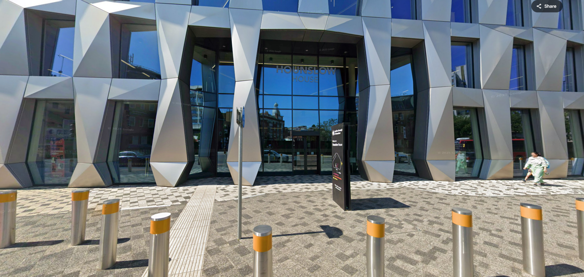 Part of a large, shiny grey building with large glass windows. The main entrance is shown, with writing reading 'Hounslow House' above the doors. A black information sign is in front of the entrance on a pavement area, with silver and yellow bollards protecting the building from the road. A woman is walking in front of the building on the right.