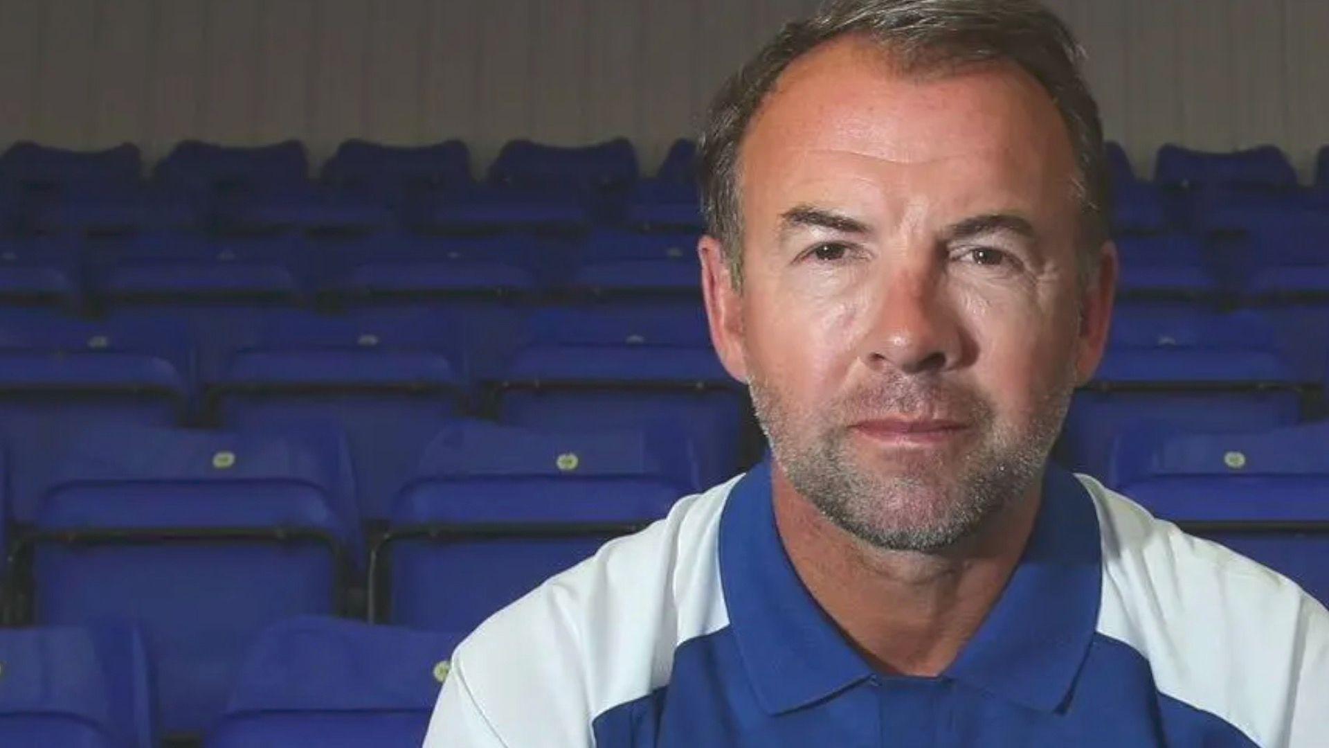 Marcus Stewart sits in an empty football stadium on a blue seat and is pictured in a blue and white Ipswich Town shirt.