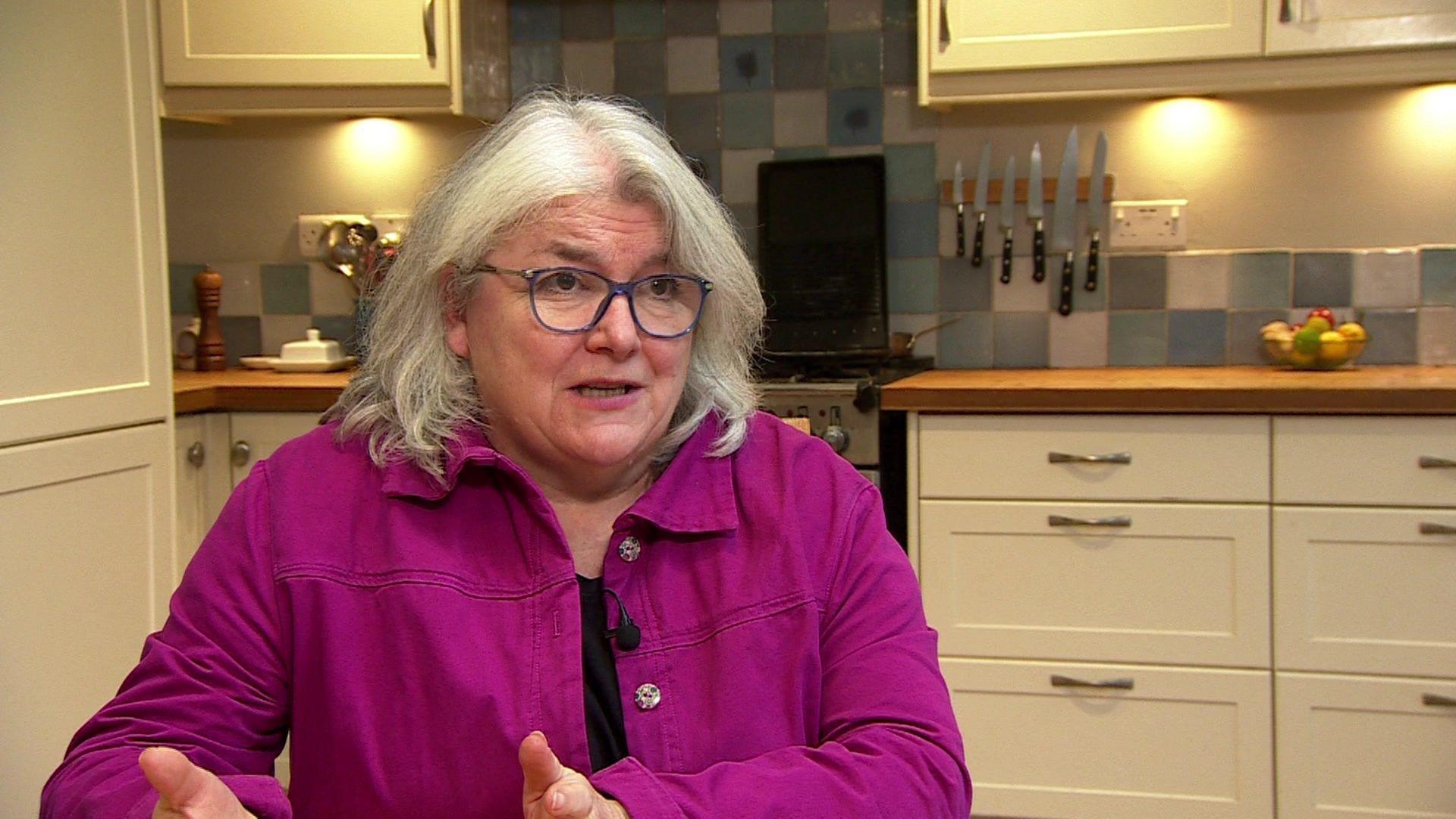 A woman with white hair wearing glasses and a pink denim jacket, talking to someone off-camera and gesturing with her hands. She is sitting in a kitchen, with counters, an oven and a tiled wall behind her.