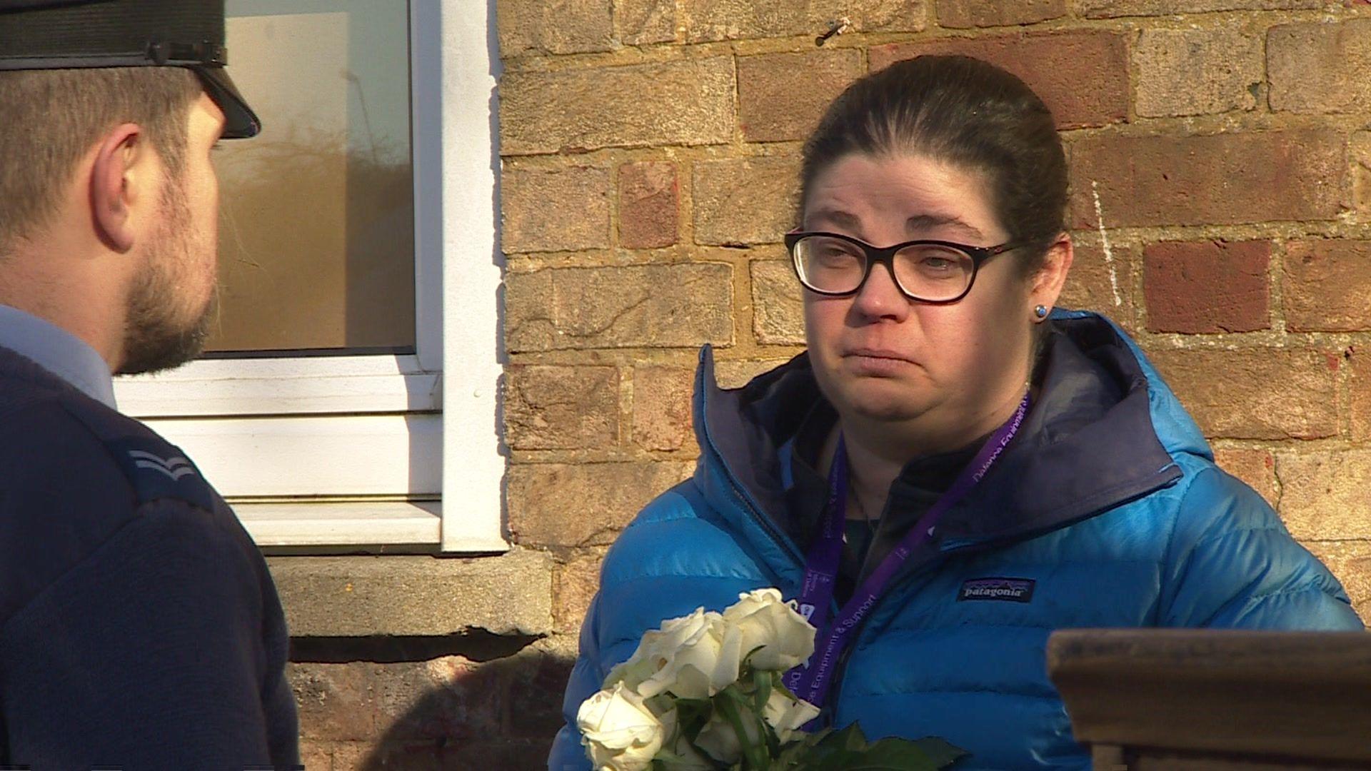 Hayley Woodhall has brown hair, glasses and is wearing a coat. She is holding white flowers in her hands.