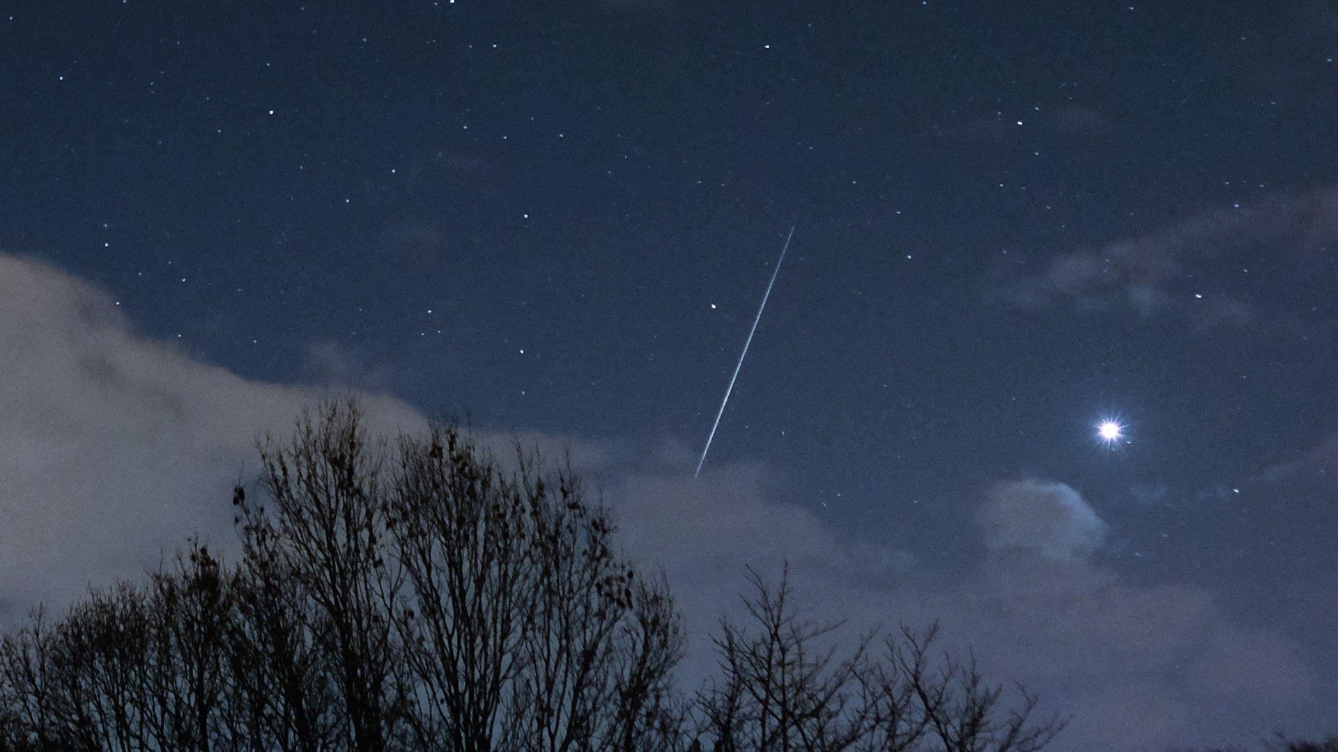 A long streak from a shooting star in a dark, clear night sky with some cloud in the foreground