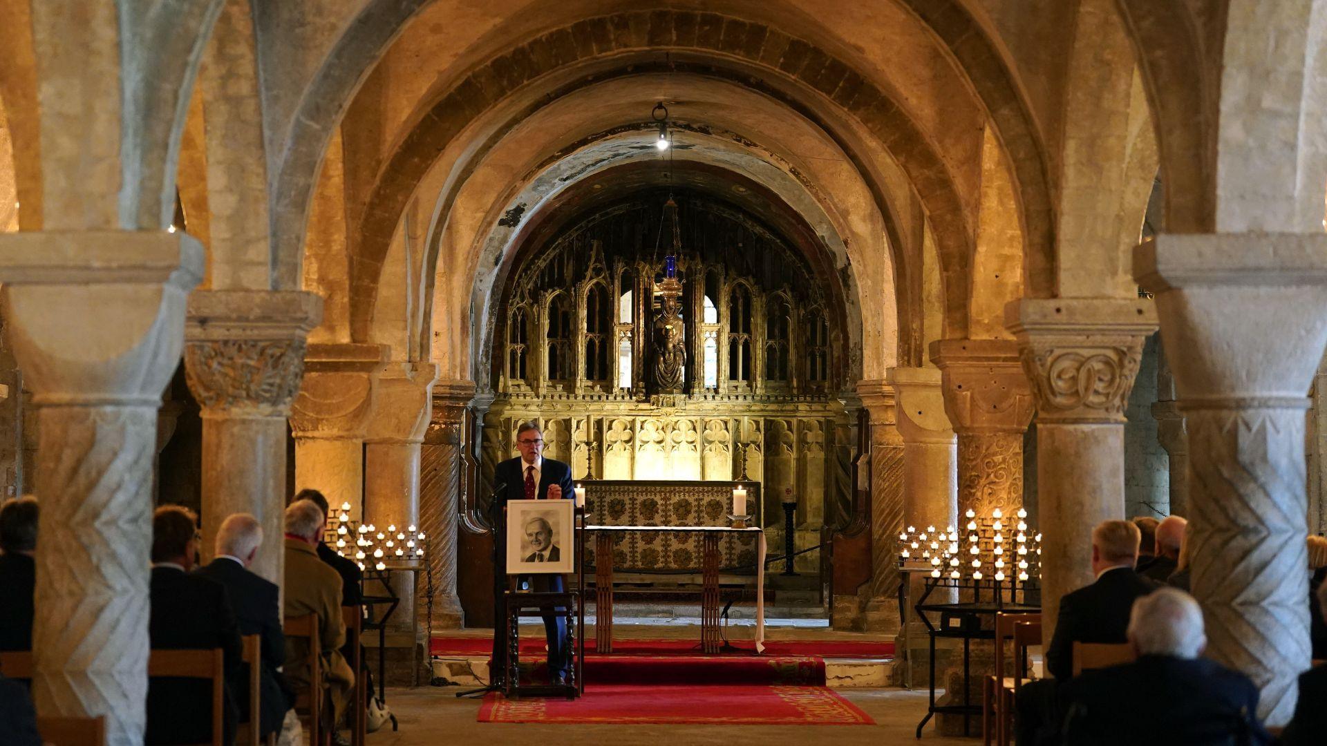 A man stood at a lectern at the front of a church while people sit facing him on chairs either side of the aisel