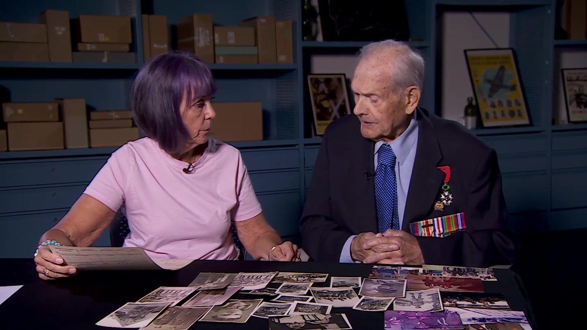 Percy, an elderly man in a light blue shirt and tie and navy blue jacket featuring military medals. He is sitting with Jacqueline, a woman wearing a pink T-shirt with purple hair. They are sitting in a blue room at the National Archives with framed historical posters behind them and a pile of photos on a black table in front of them.