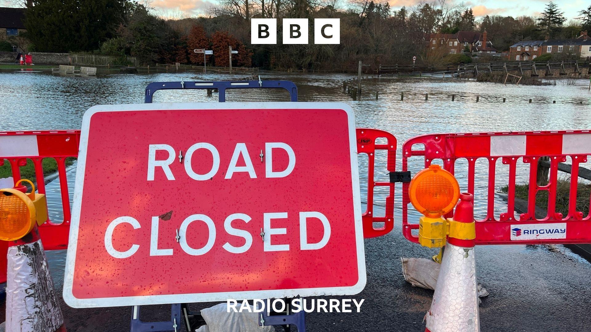 A road closed sign stands in front of a road and village green both completely submerged under water. 