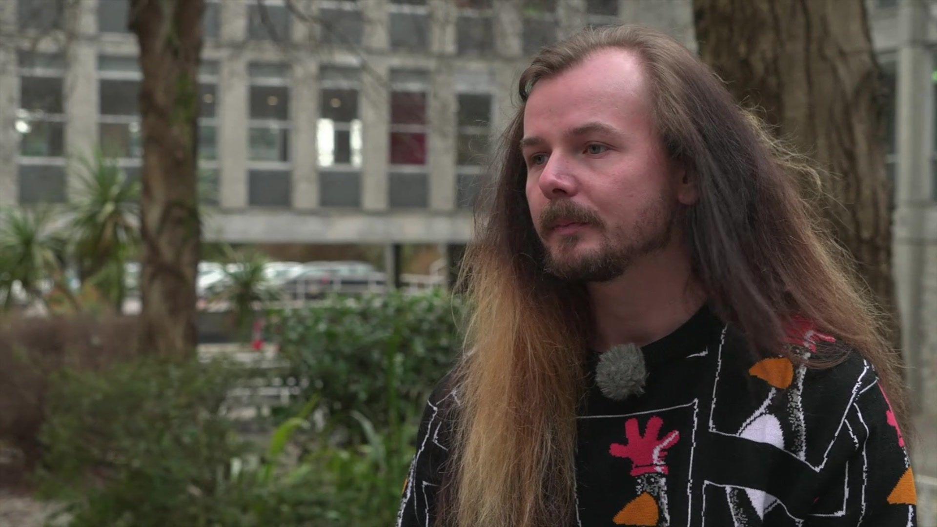 Jack Wilkes is seen outdoors here, next to trees with a grey office-type building behind him. He has long hair, a beard and a moustache, and is wearing a black sweater with pictures of the Wallace and Gromit penguin character Feathers McGraw on it. 