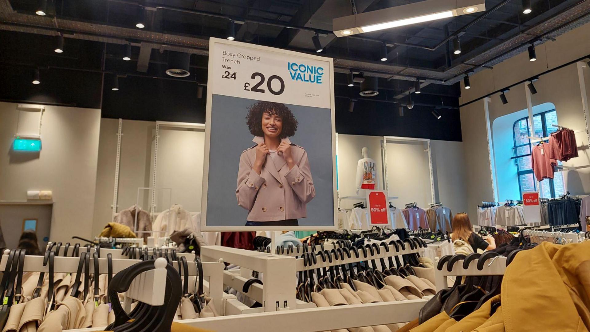 Clothing rails filled with beige and mustard trench coats in the foreground of a clothing shop. A large promotional sign above the display shows a cropped trench coat priced at £20, reduced from £24, with the text “ICONIC VALUE”. Additional clothing racks extend across the shop floor, including tops and jackets, with red sale signs showing “50% off”. A mannequin stands near the back of the store, and overhead ceiling lights illuminate the retail space.