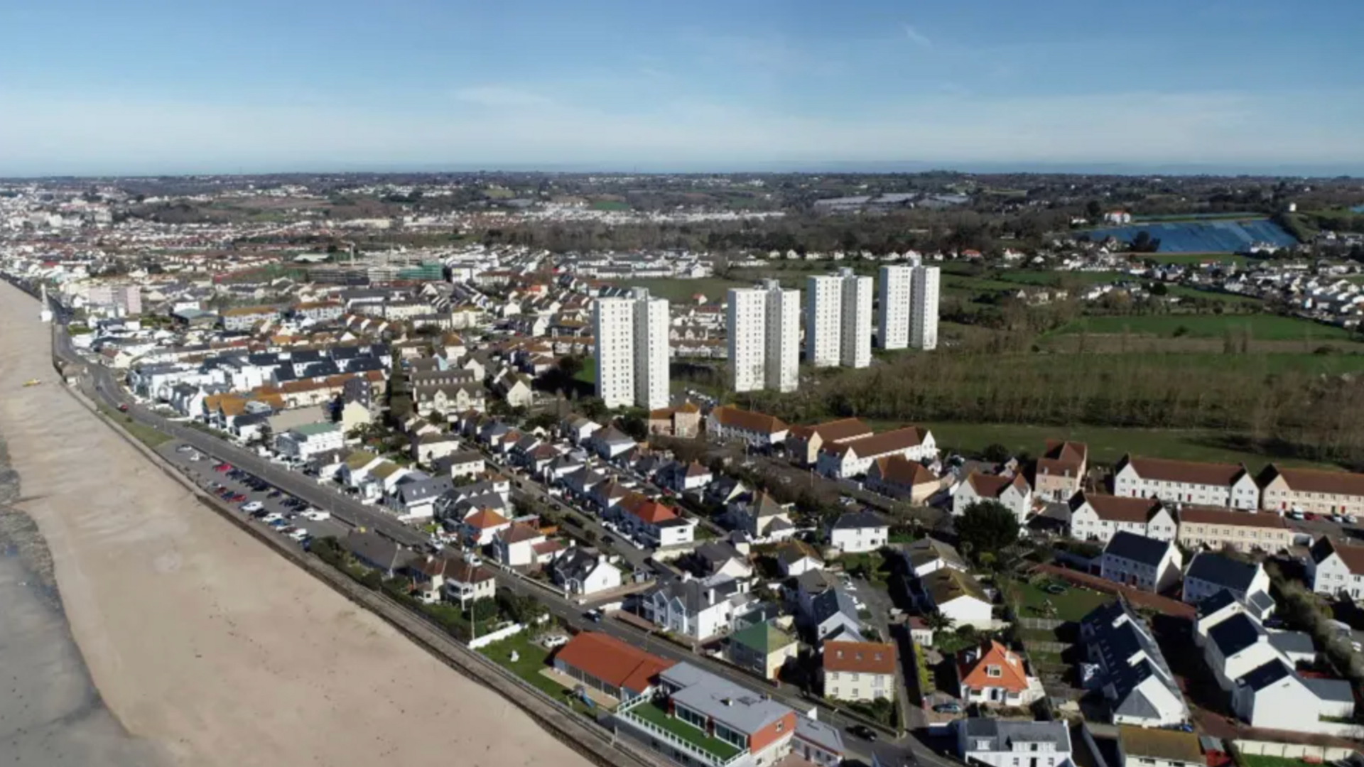 An aerial view of Jersey with the beach in view and the main road followed by rows of houses and four large blocks of flats, blue skies