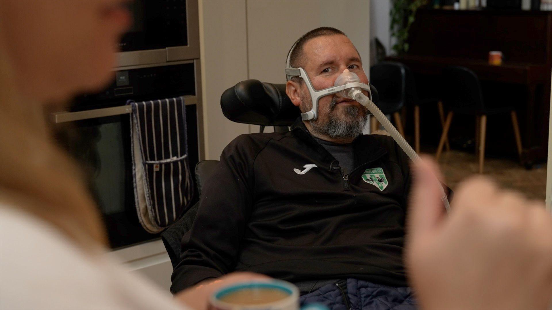 Nik Ward, an MND sufferer, sitting in his motorised wheelchair while he looks towards a family member standing to the left of the camera. He is dressed in a black tracksuit jacket and is using a breathing apparatus across his nose. 