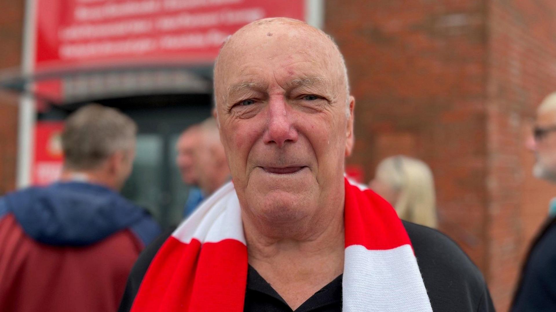 Tony Williams wearing a Wrexham red and white striped football club scarf untied around his shoulders, smiling into the camera stood in front of people entering the Wrexham AFC grounds.