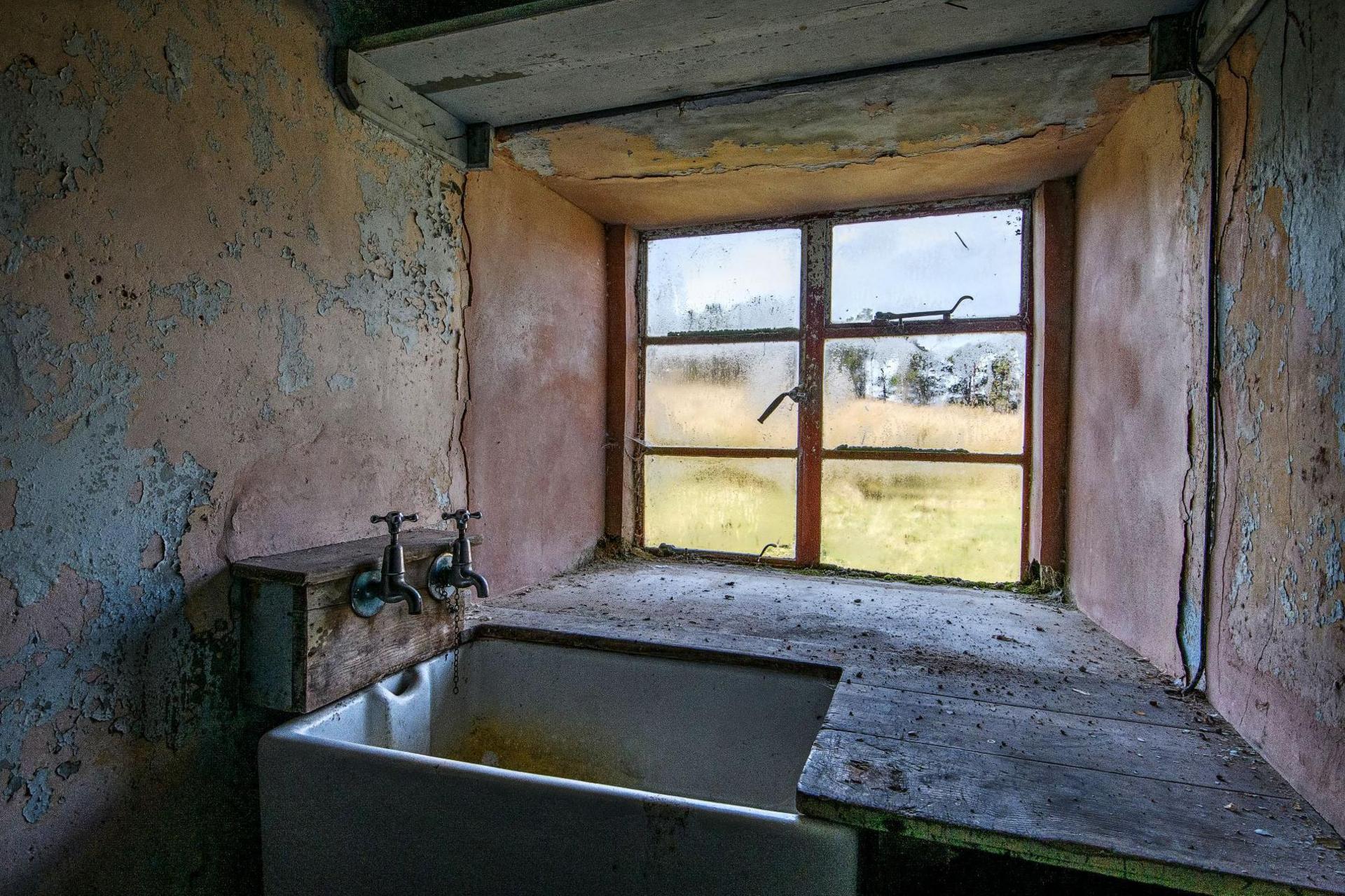 There is a small window above a small, stone sink. Two taps stick out from a wooden box above the sink.