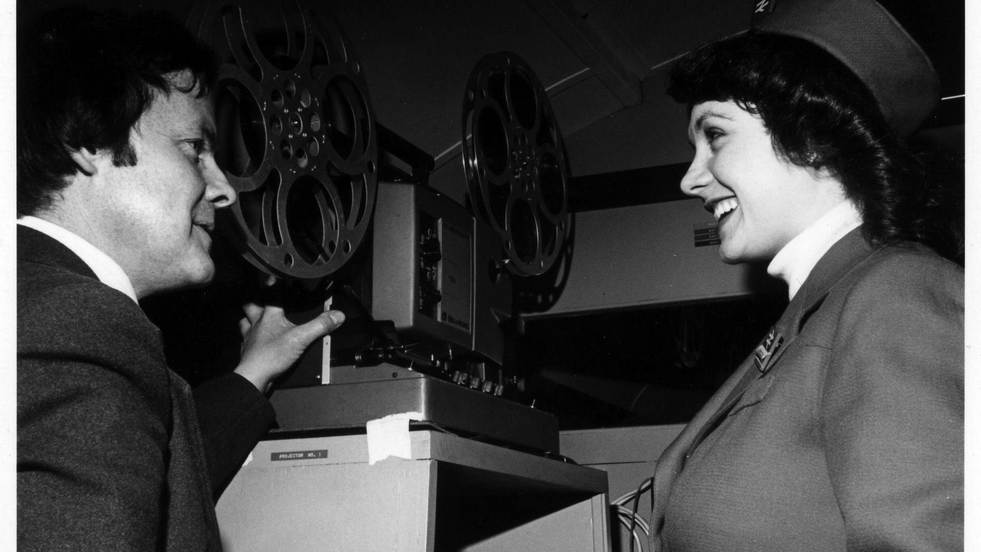 An old black and white photo showing Alan Willmott next to a large reel projector speaking to a woman, who is smiling at him. They are both smartly dressed in suits.