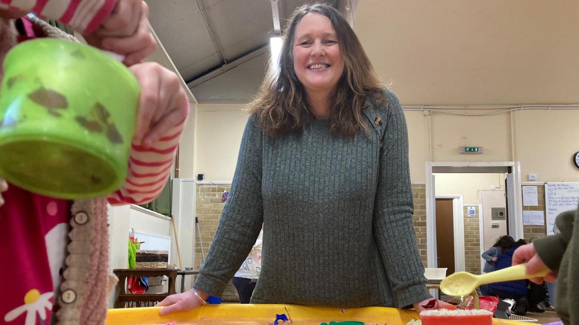 A smiley woman with long dark hair wearing a green jumper stands infront of a low children's table where small children's hands can be seen playing with buckets and spades