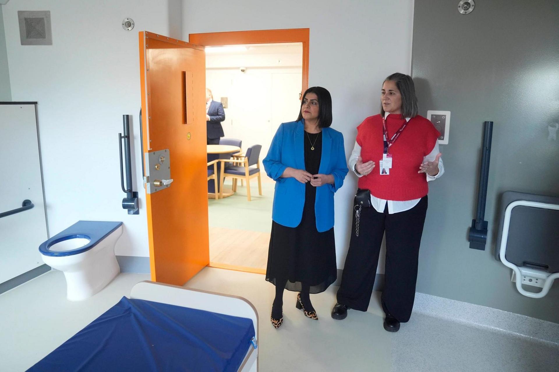 Two women stand inside a cell in a prison, which has a bed, toilet and what appears to be a pull-down table. The woman on the left has black, shoulder-length hair and wears a blue jacket over black dress. The woman on the right has brown shoulder-length hair and wears a red jumper over a white blouse and black trousers. The cell door is orange and the bed cover is blue.