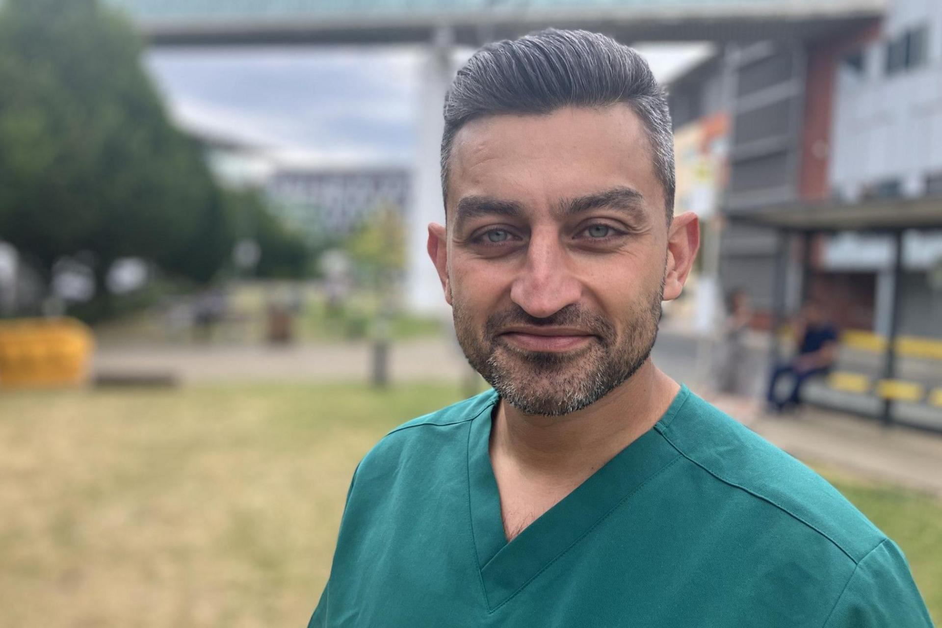 The image shows a close-up portrait of a person standing outdoors, wearing teal V-neck medical scrubs. The photograph is sharply focused on the person in the foreground, while the background is blurred with a shallow depth-of-field effect. In the background, there appears to be a grassy area, trees, a modern building, and a bus stop or shelter with a few people nearby. 