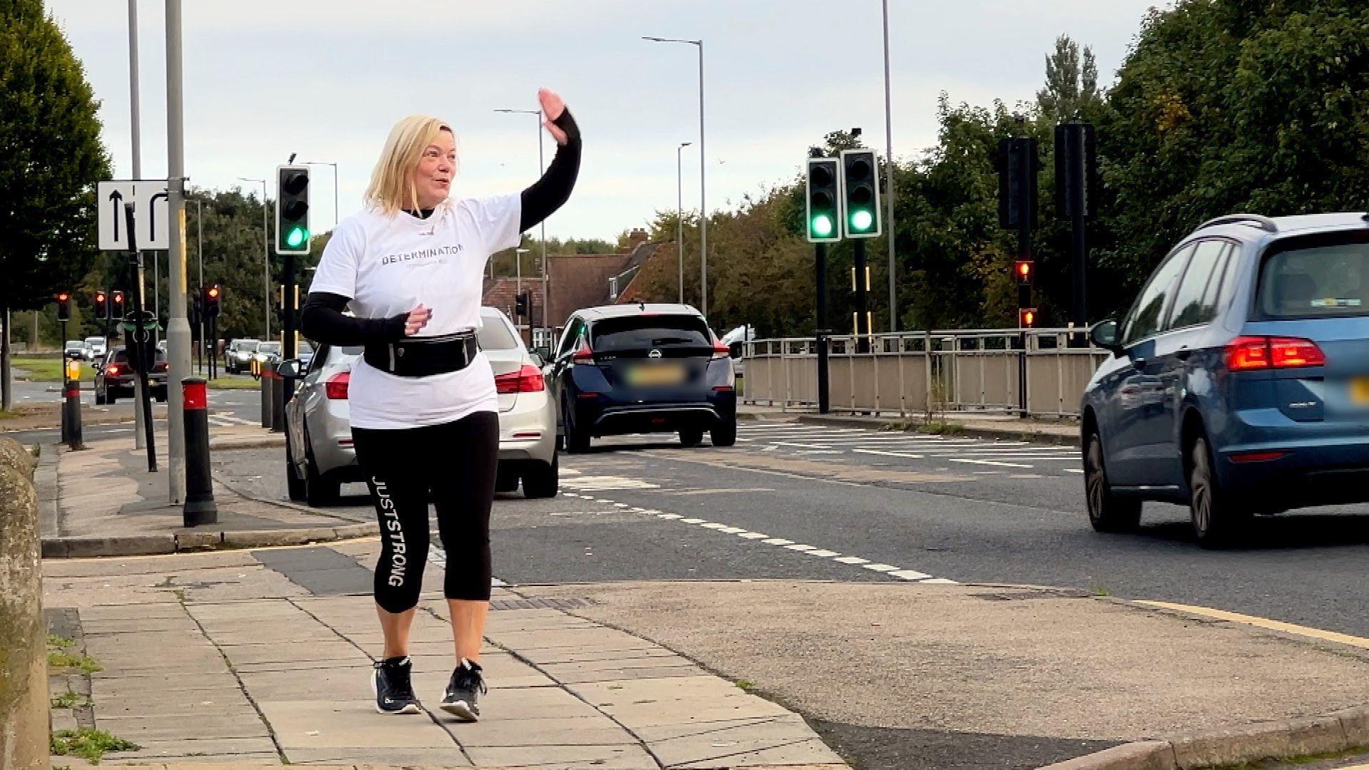 A woman walking and dancing alongside a busy road. She is wearing a white short-sleeved top over a black long-sleeved one and slightly cropped black sports leggings. She is waving to someone off camera as cars drive past.