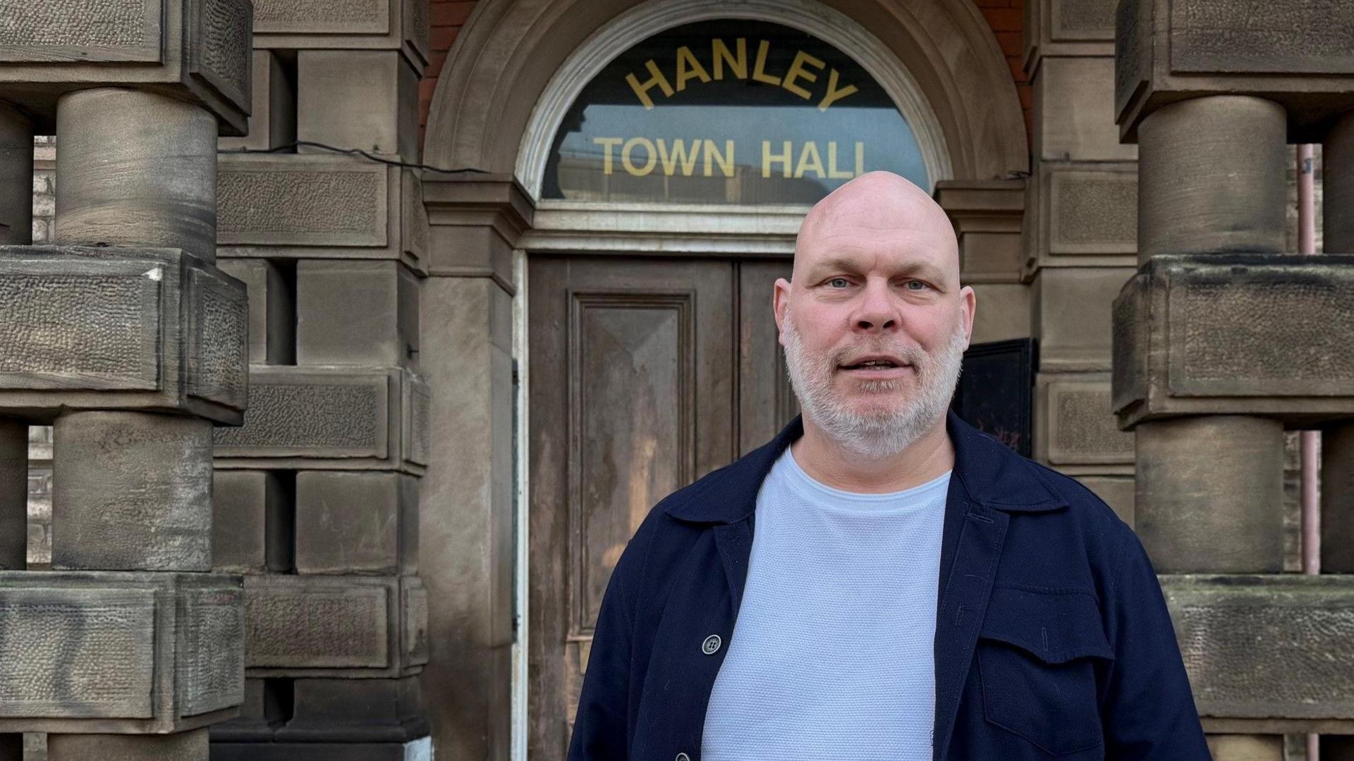 Simon Edwards, a bald man with a grey beard, wearing a navy blue jacket over a white t-shirt. He is standing outside a stone building which has the words Hanley Town Hall in gold lettering on a arched window above a wooden door.