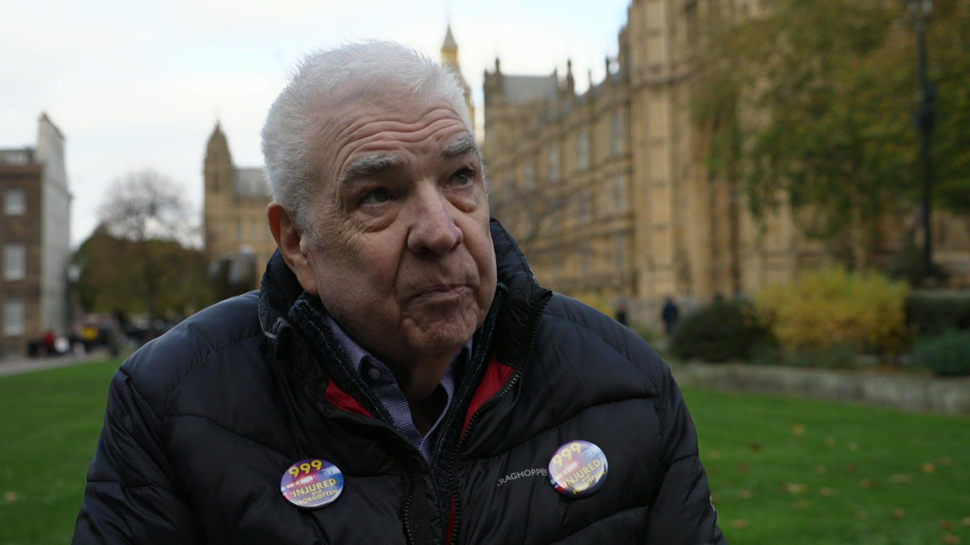 Tom Curry - a man wearing a black coat with badges on reading "999 INJURED AND FORGOTTEN". He is standoing outside the Houses of Parliament.