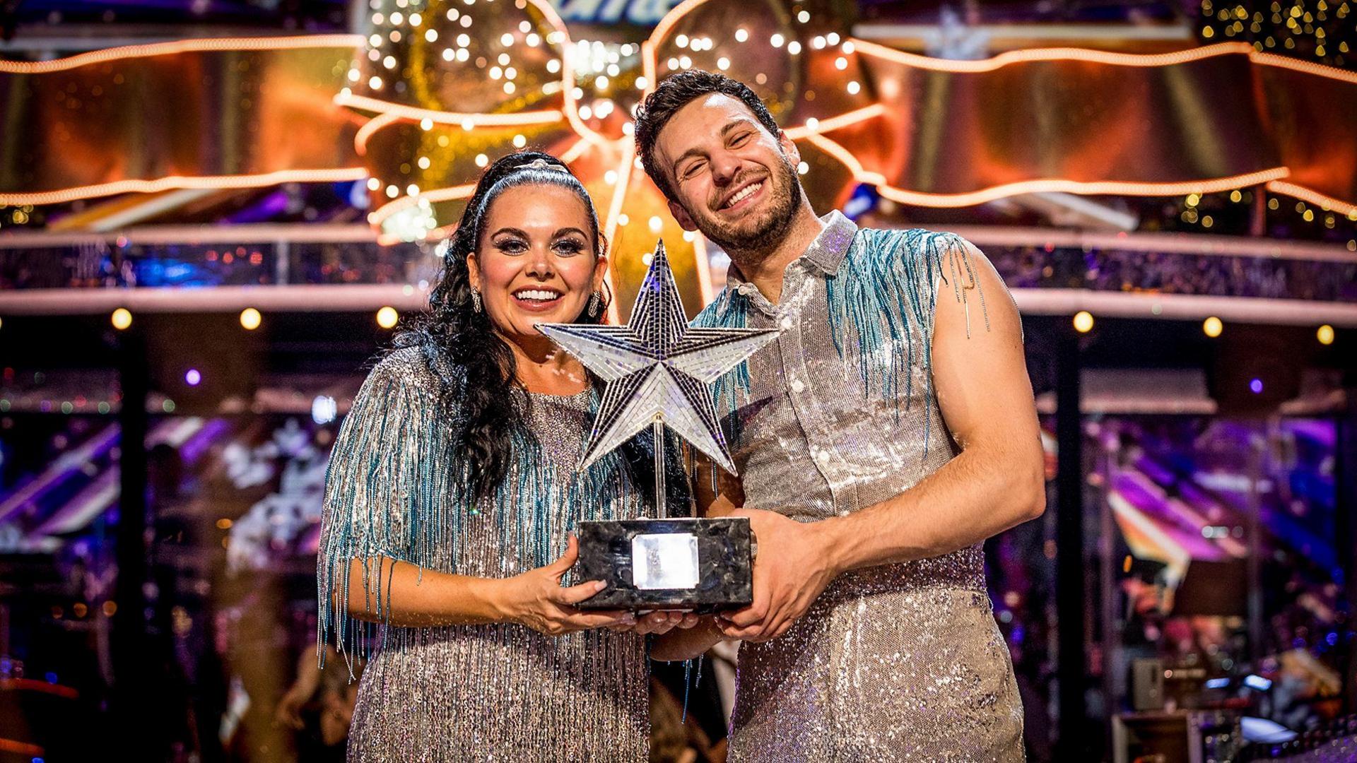 Scarlett Moffatt and her professional dancer smile as they hold a trophy with a silver star on