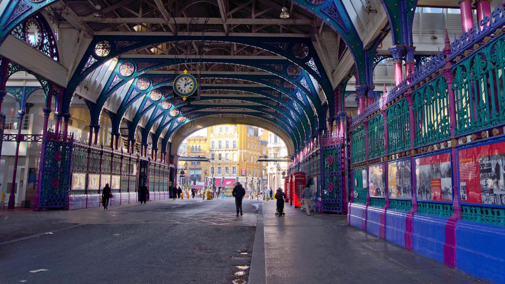 The market is empty, with a few people in the distance walking though the open space. The market opens out onto buildings and on both sides are intricate iron railings painted purple, green and blue. 