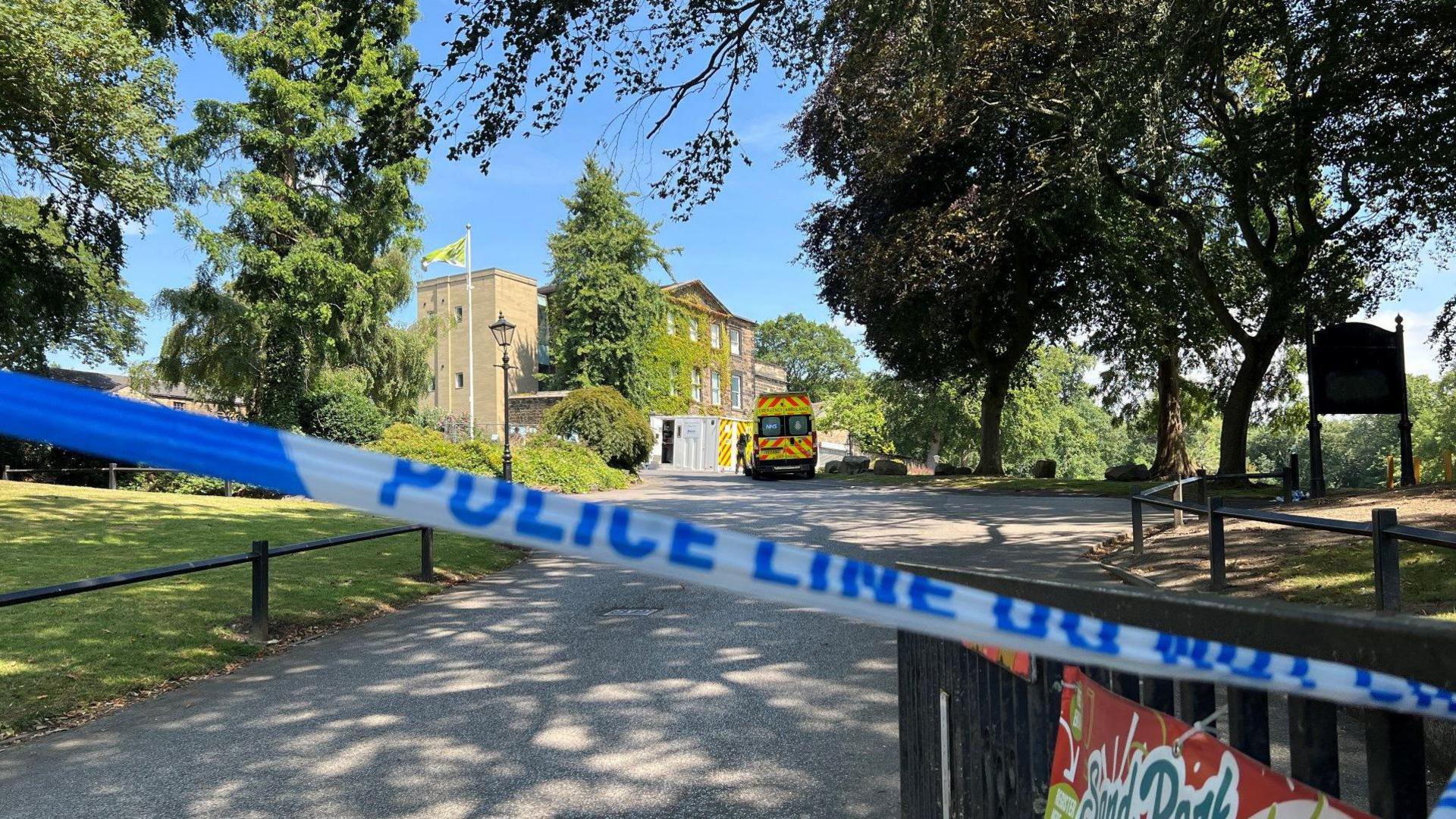 A line on blue and white police tape hangs across the front of the entrance to Crow Nest park. In the background a police van can be seen parked outside a large building. There are trees and grass either side of a path leading up to the building.