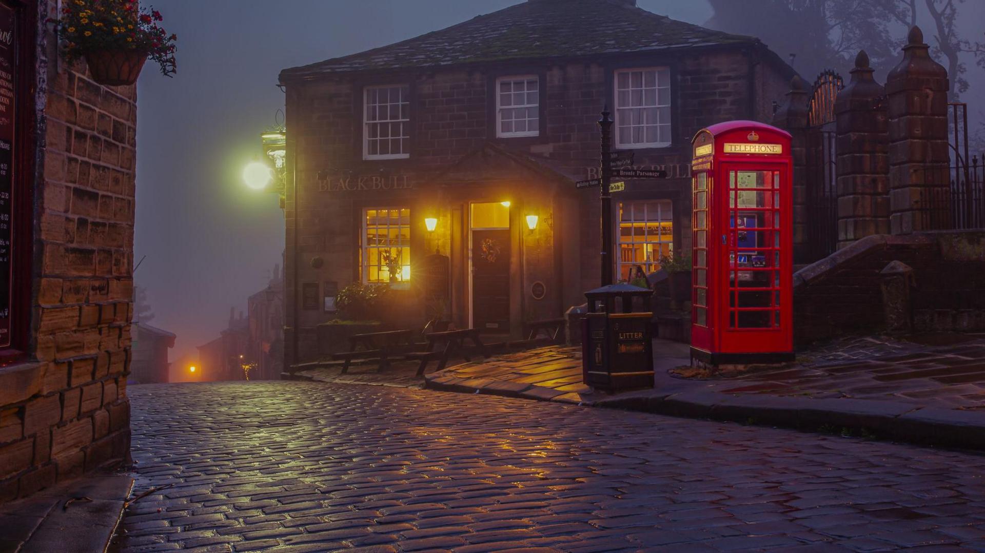 A stone-built pub in twilight, with a sign saying "The Black Bull", is in the background, lit up by yellow lantern-light, while in the foreground is a traditional red telephone box, a black and gold litter bin and a wet cobbled street reflecting streetlights.