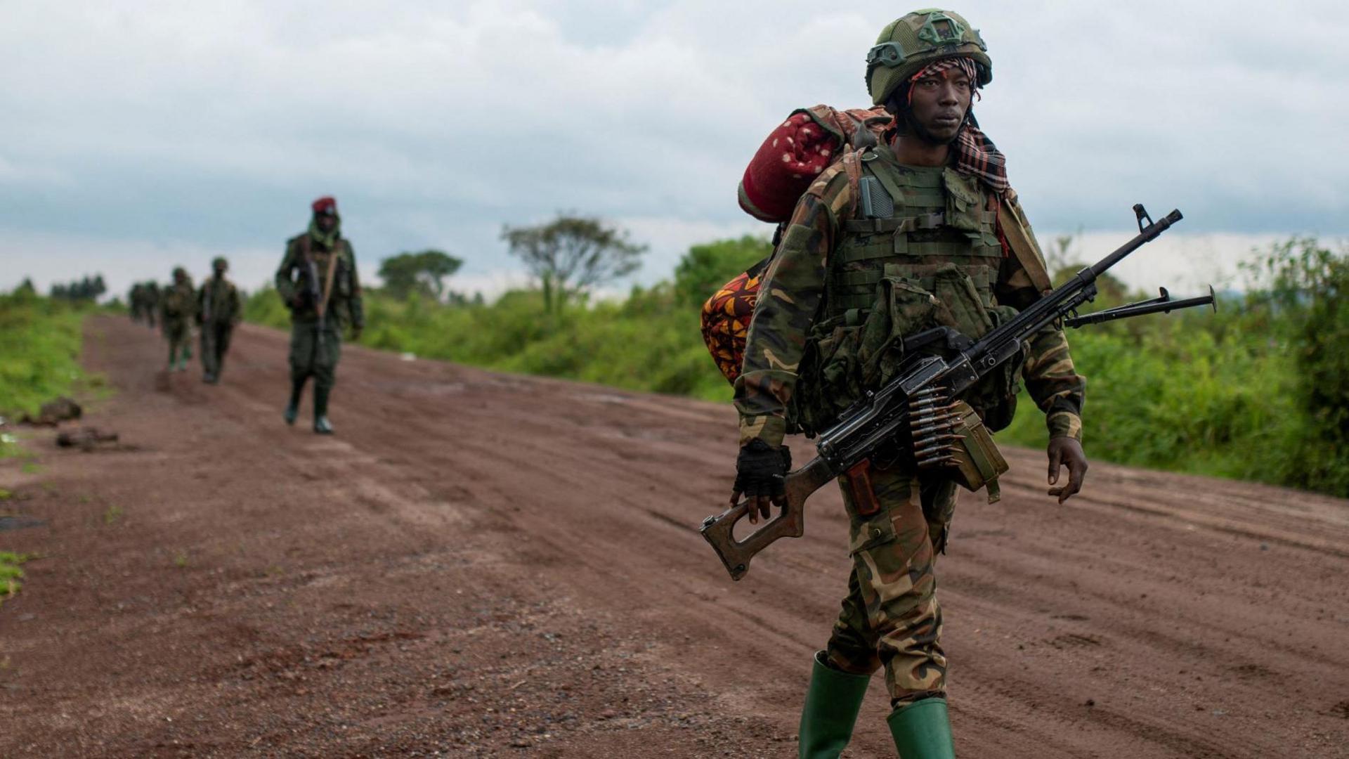 An M23 fighting in camouflage uniform walking along a dirt road in DR Congo, followed by his comrades. He is holding a gun, and carrying a large backpack.