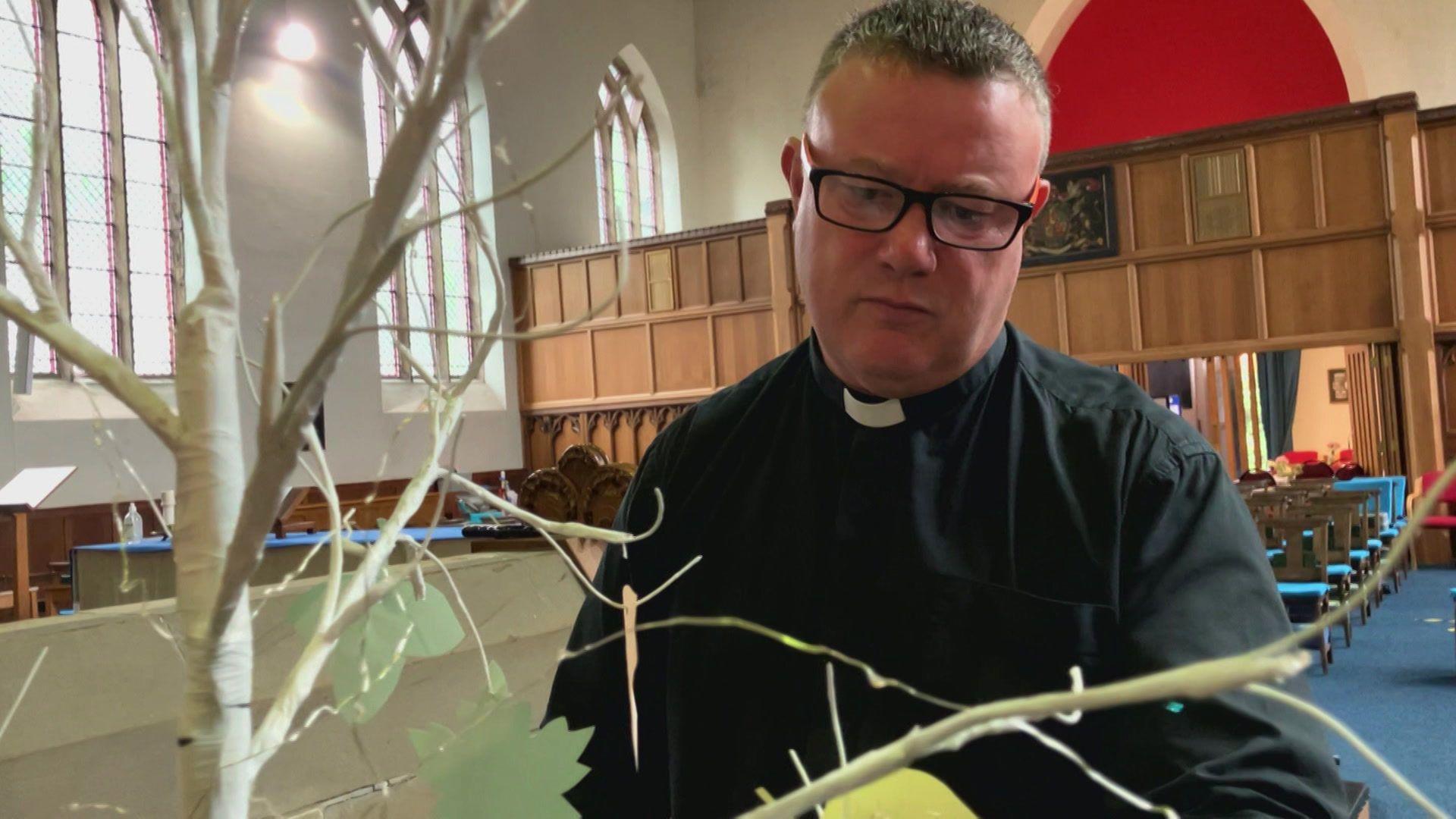 A man with short hair and wearing glasses looks at small pieces of paper hanging from a tree-like structure, which is painted white.
