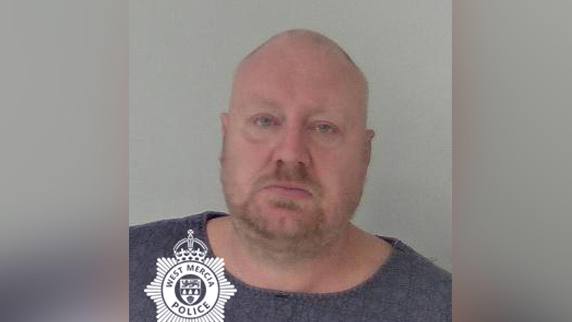 Police mugshot showing a bald man with short facial stubble  wearing a blue T-shirt and looking at the camera