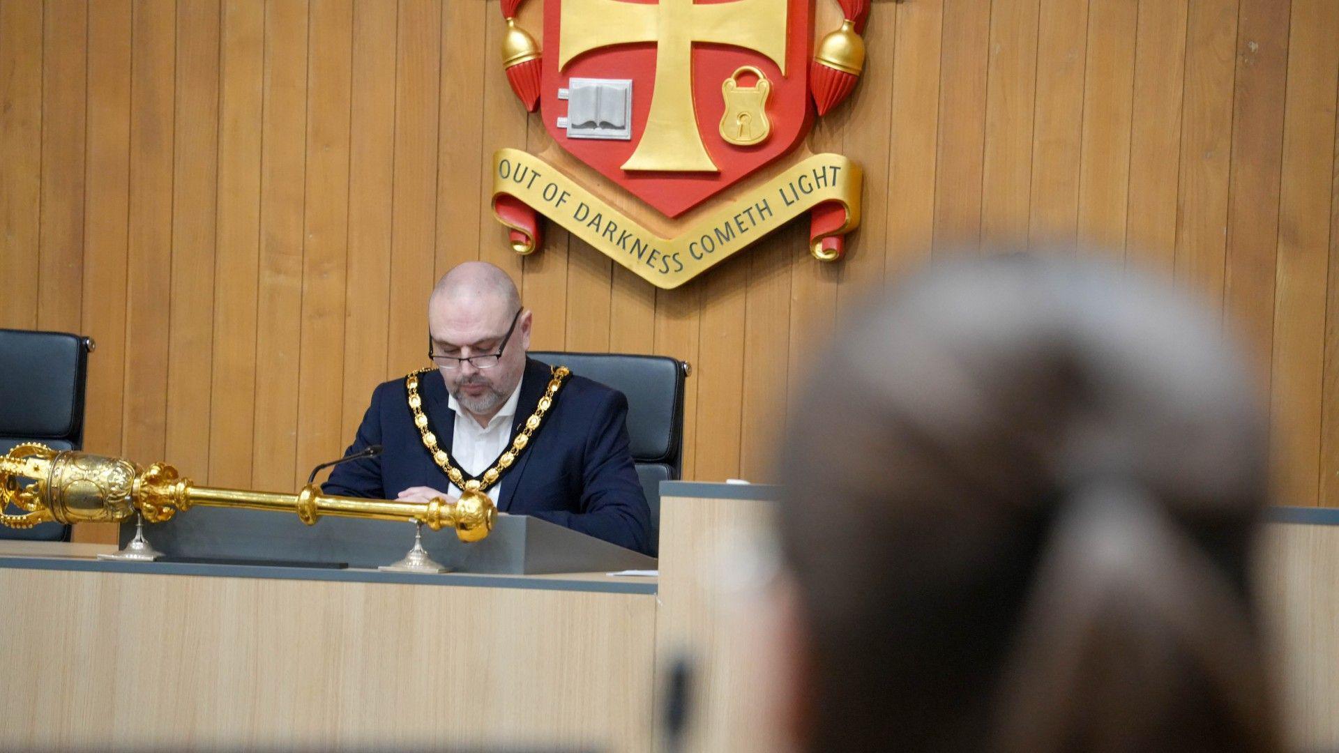 The deputy mayor underneath a crest of Wolverhampton. There is a ceremonial baton next to him and he is wearing jewels around his neck. 