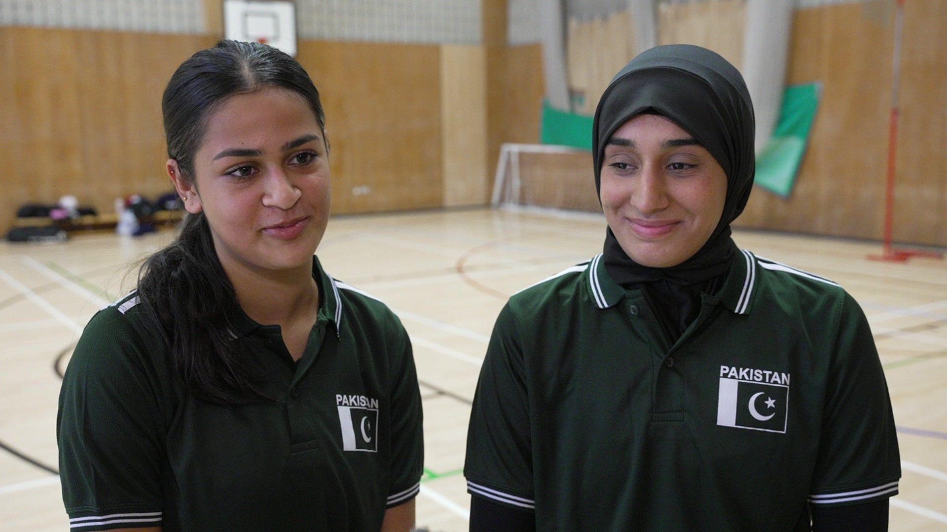 Sarina has long dark hair pulled back from her face in a ponytail. She is wearing a black polo top with the Pakistan team logo top left. Safeeyah stands to her left and wears a black hijab and a black polo top with the Pakistan logo top left