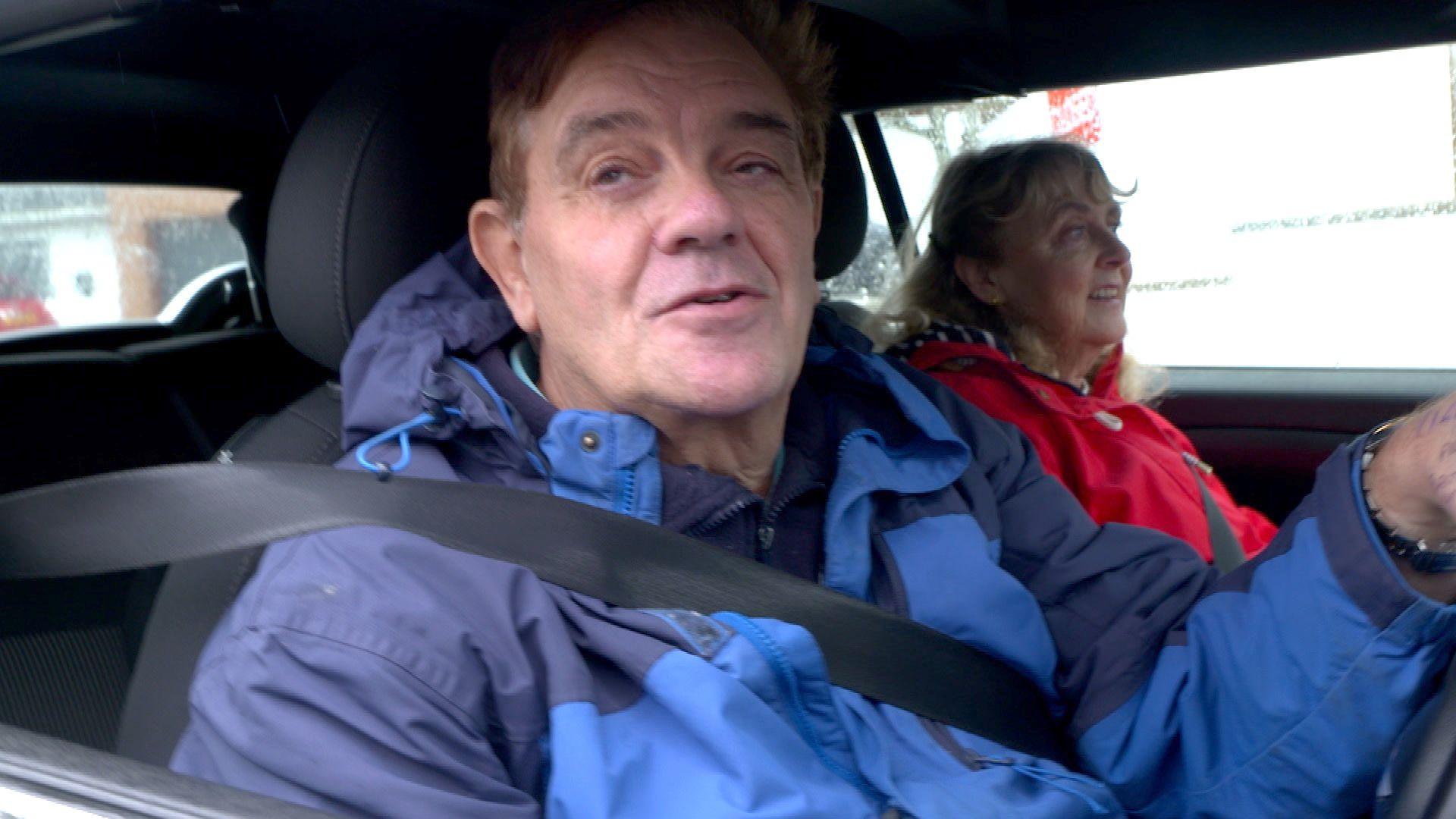 Peter Shirreff inside his vehicle wearing a dark and light blue coloured jacket. He is wearing his seatbelt and a woman, wearing a red coat, is sat in the passenger seat. He has one hand on the steering wheel.