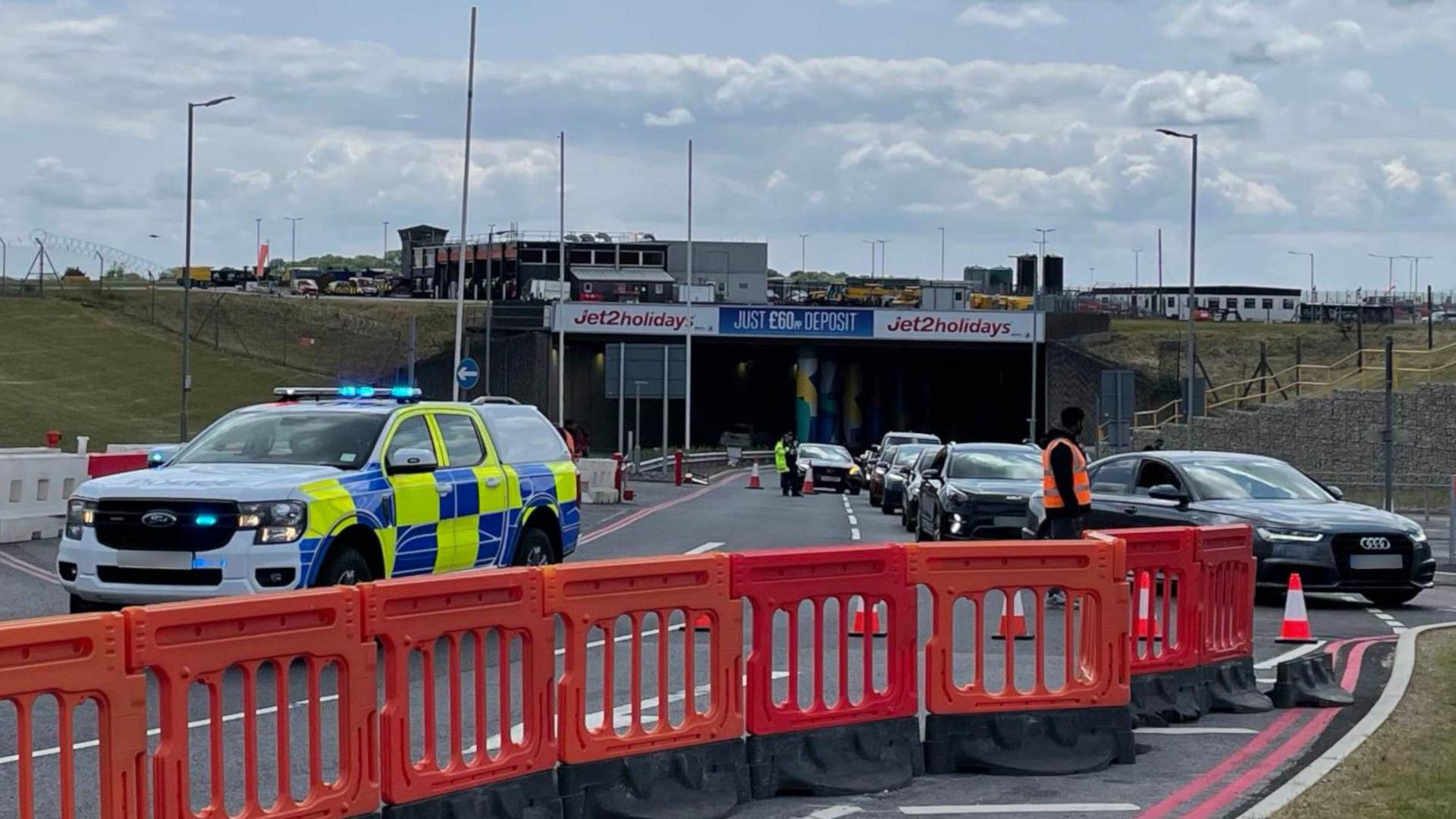 A police truck has its blue lights on behind a line of orange barriers shutting the road. It is blocking the entrance to a tunnel at the airport, which has a stream of cars queuing to get out.