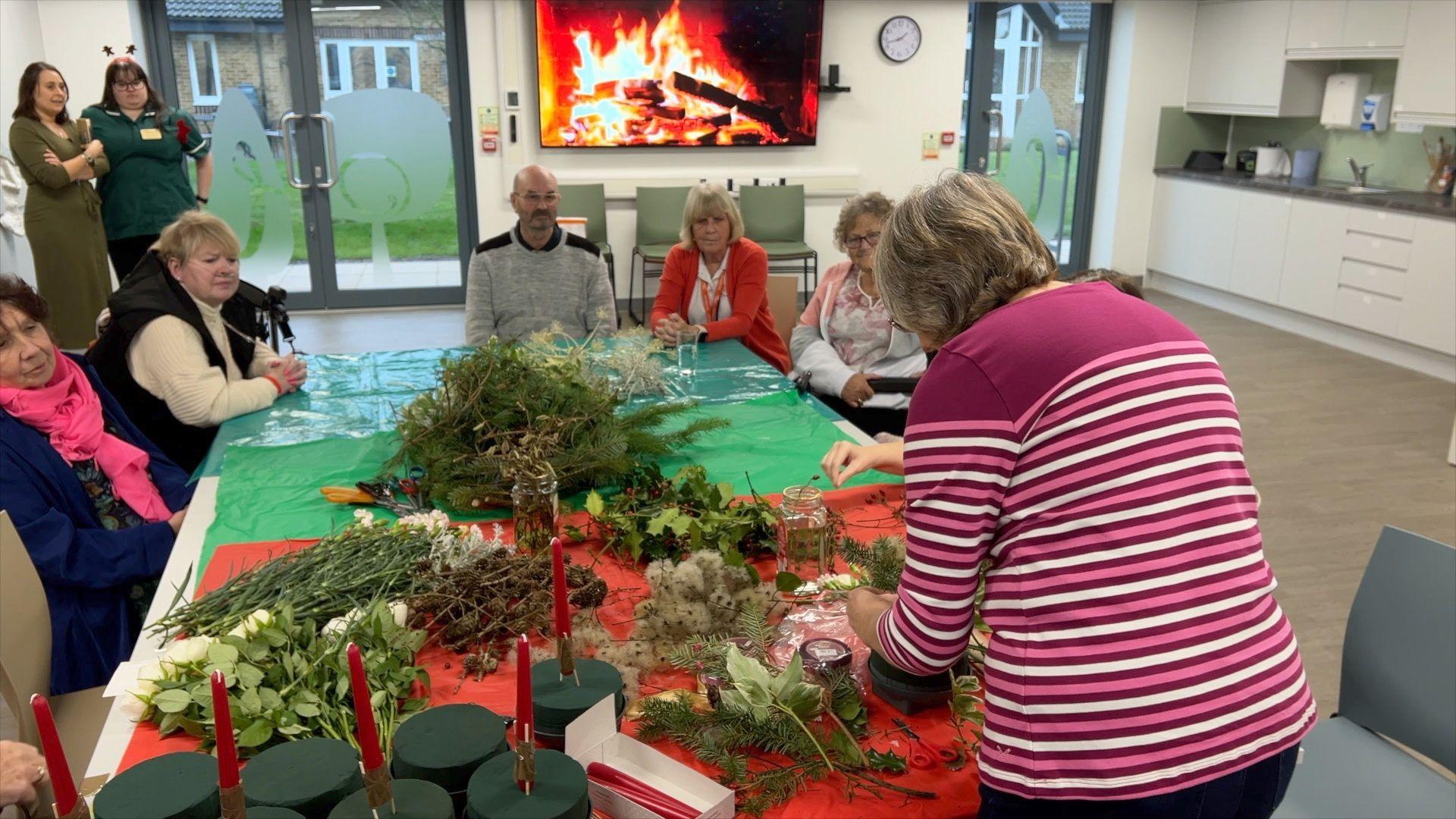 Five people including hospice patients are seated at a large table facing the camera, watching a teacher show them how to make Christmas table decorations 