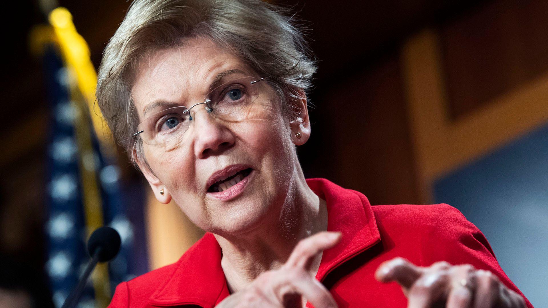 Sen Elizabeth Warren, with short grey hair and a red suit, speaking at a press conference 