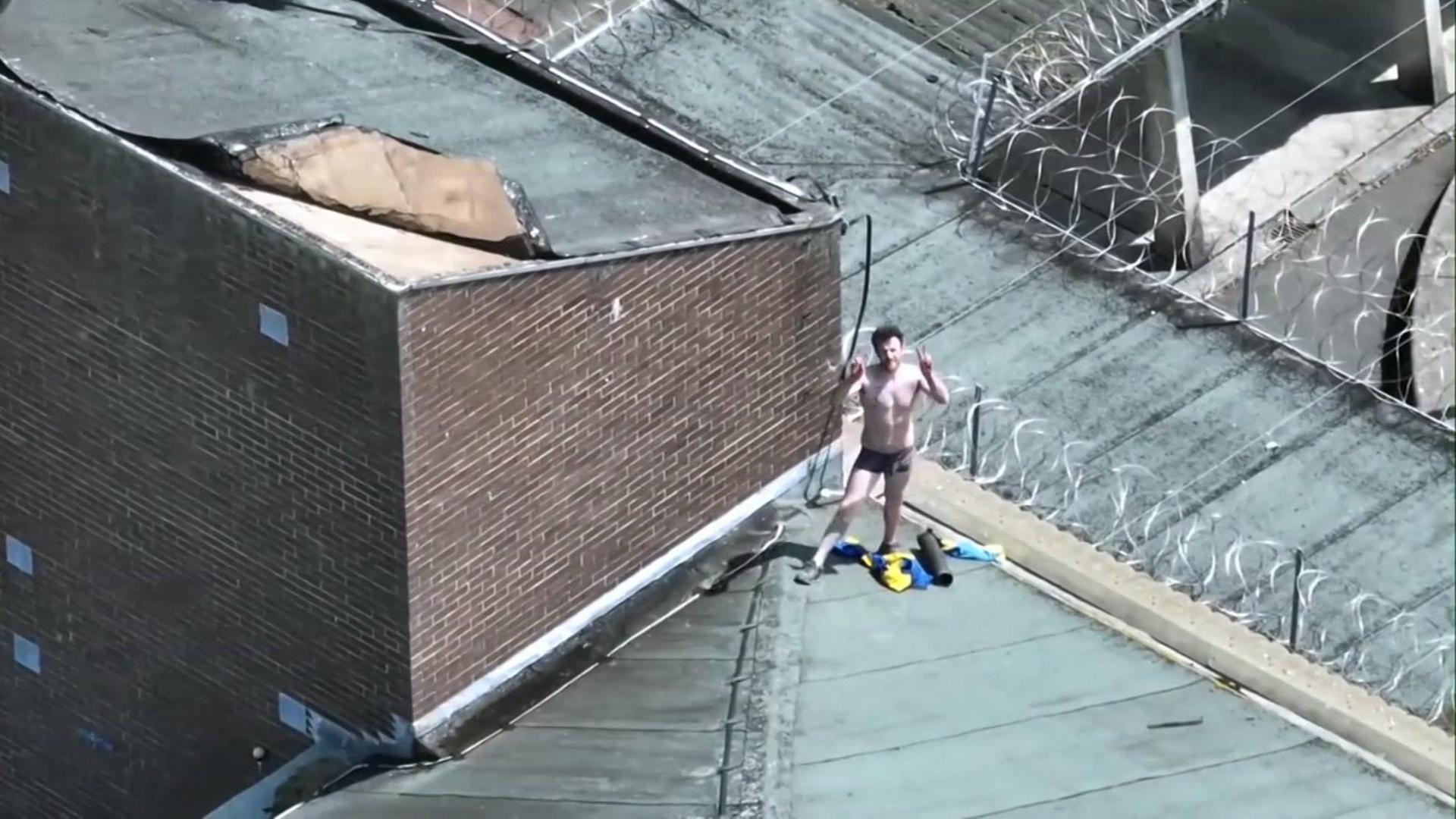 Joe Outlaw, wearing just black boxer shorts, stands on top of a prison roof making peace signs with his fingers. 