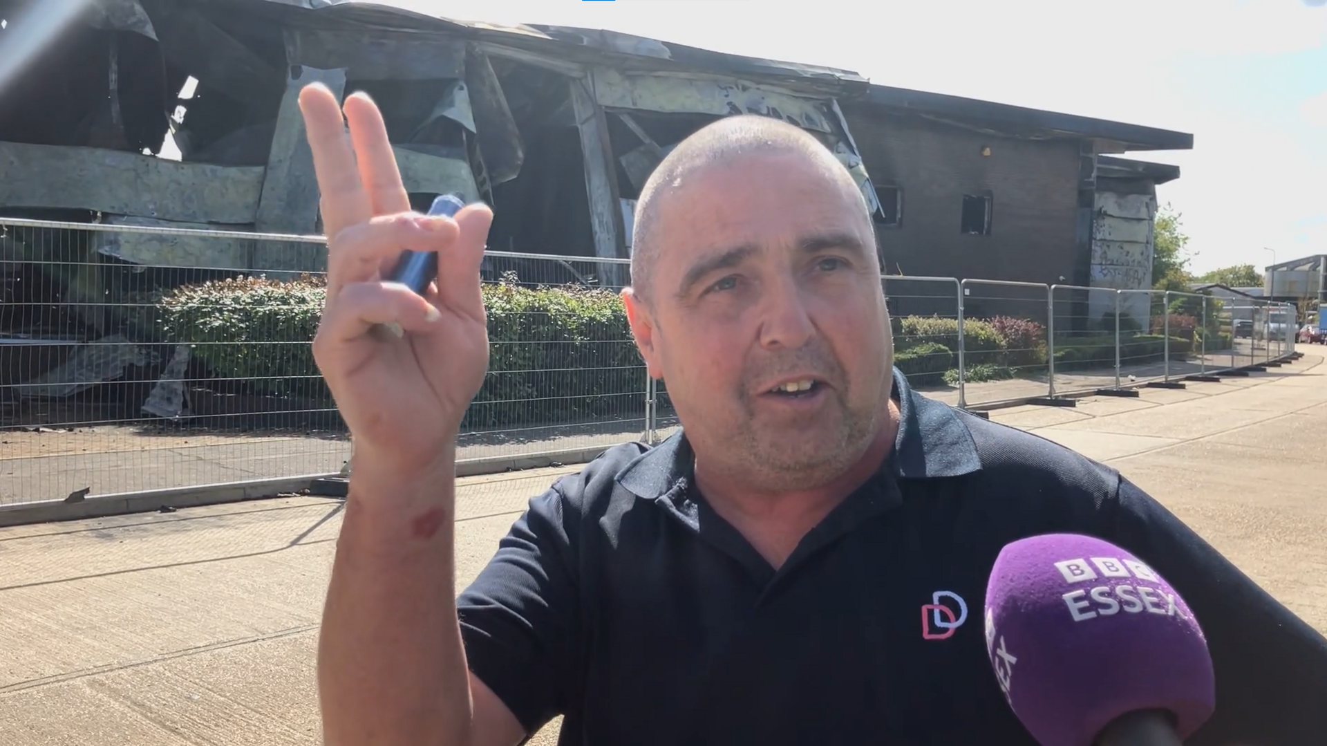 A man gesturing while talking into a BBC Essex microphone, in the background is a burnt building