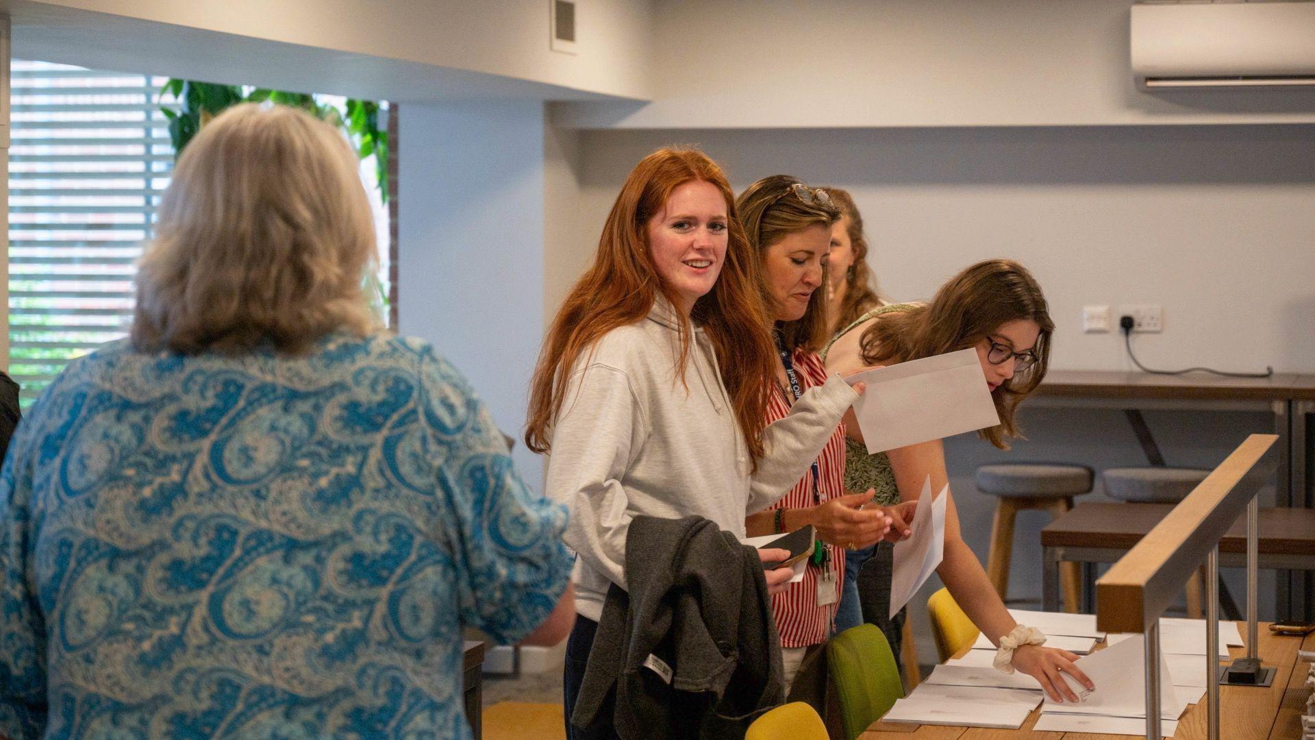 A group of young women stand by a table which has envelopes on it. They are either smiling and looking down at the envelopes or looking to the side as if talking to someone. In the centre of the image is a young woman with long red hair, wearing a light grey hoodie, she is holding an envelope and smiling.