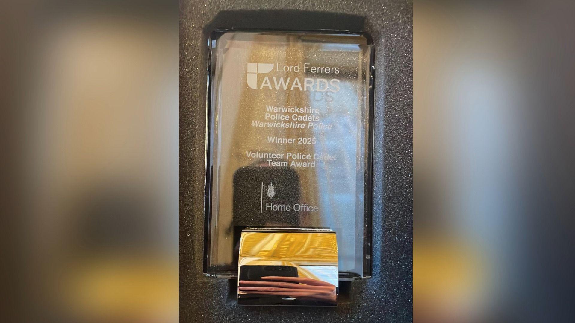 A glass trophy in a grey padded box. The inscription contains the Lord Ferrers Awards logo at the top and underneath says Warwickshire Police Cadets, Warwickshire Police, Winner 2025, Volunteer Police Cadet Team Award. The Home Office logo is inscribed below.