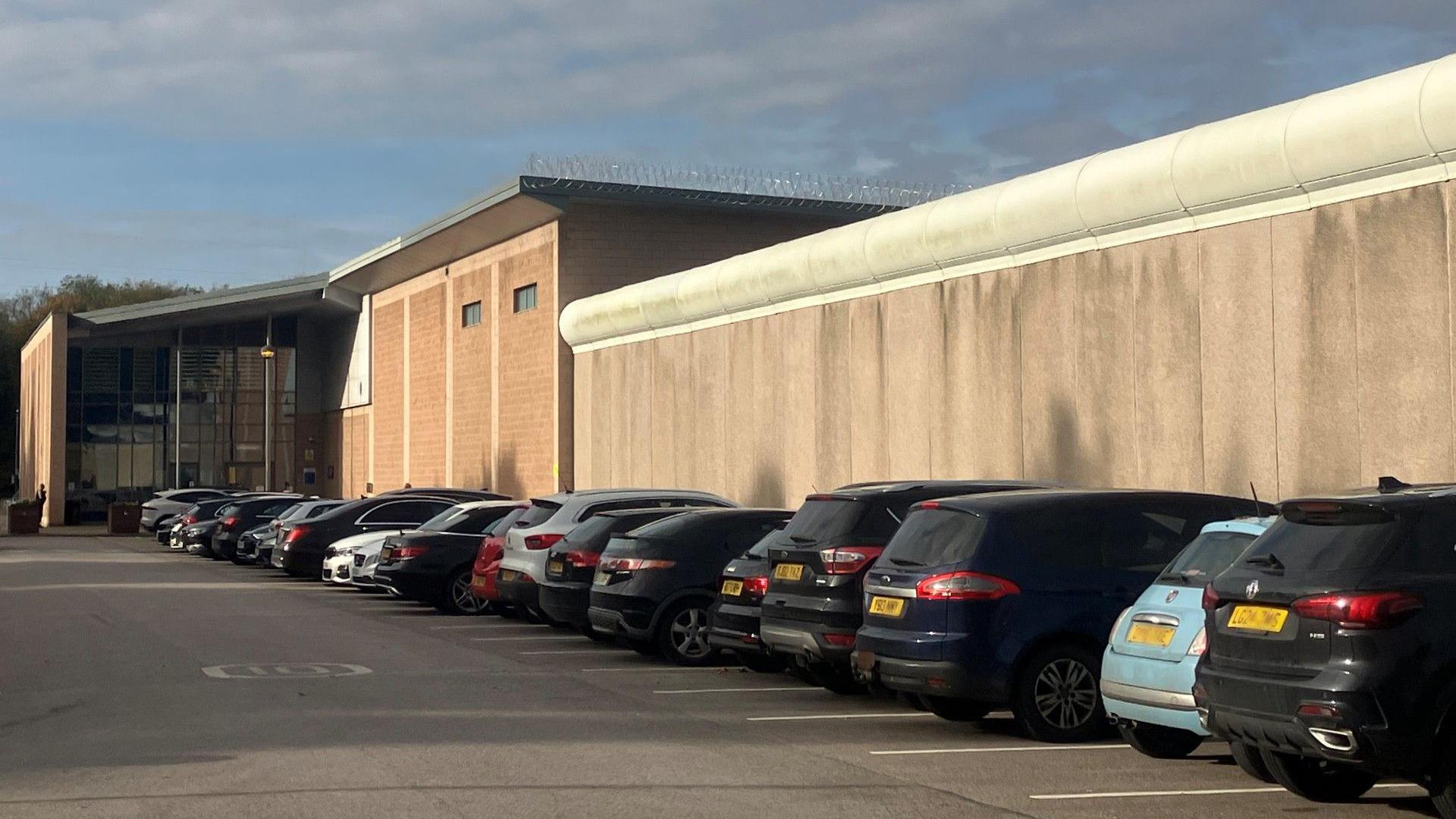 Cars parked outside a prison wall by the entrance to Forest Bank private prison, with the windowed entrance to the jail seen at the end of the car park. Barbed wire can be seen trailed along the edge of one of the rooftops. 