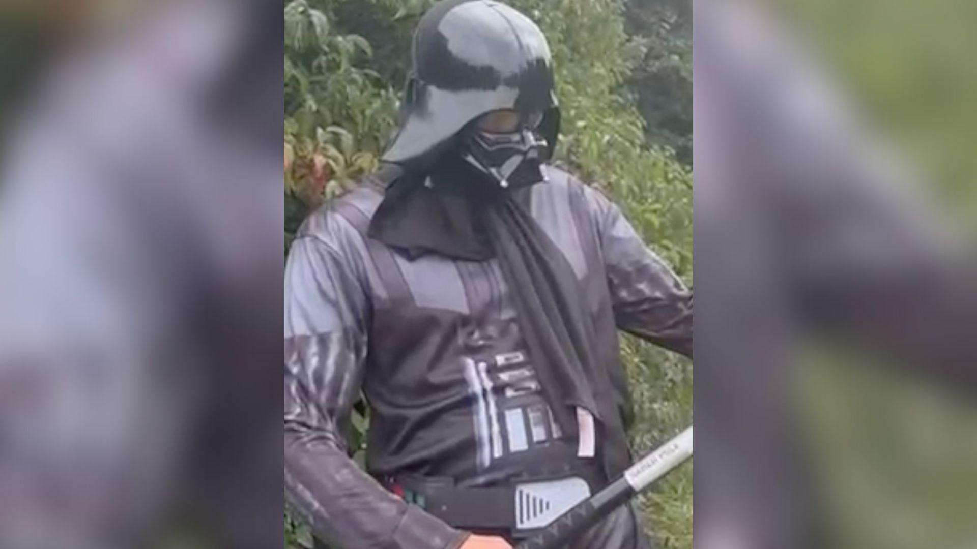 A man in a Darth Vader fancy dress costume standing in front of bushes. He is looking downwards at his light sabre. 