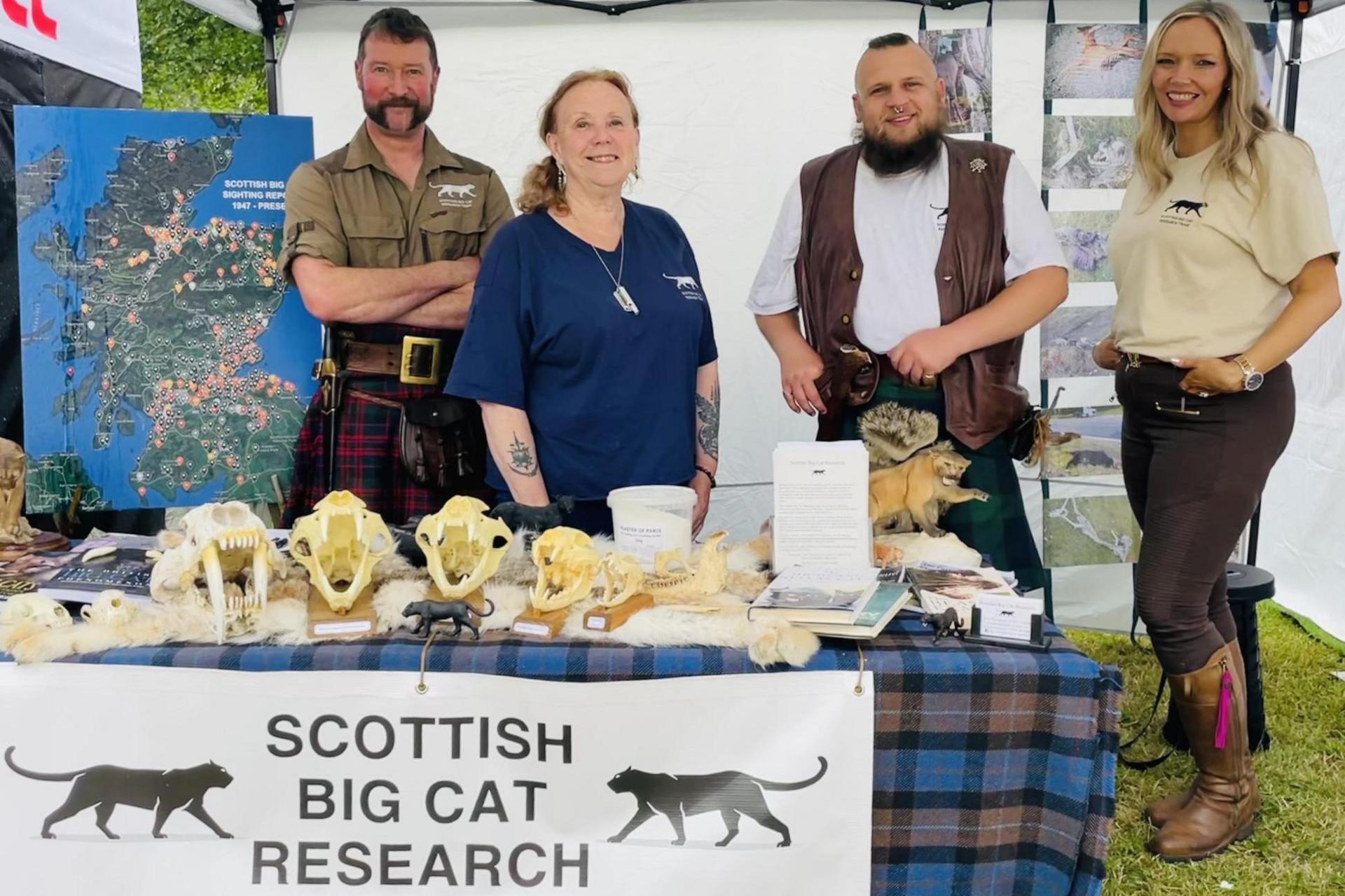 The four members of the project are at an event. They are standing next to a table draped in tartan and with a selection of big cat skulls set out on it. They also have a map showing where big cat sightings have been reported.